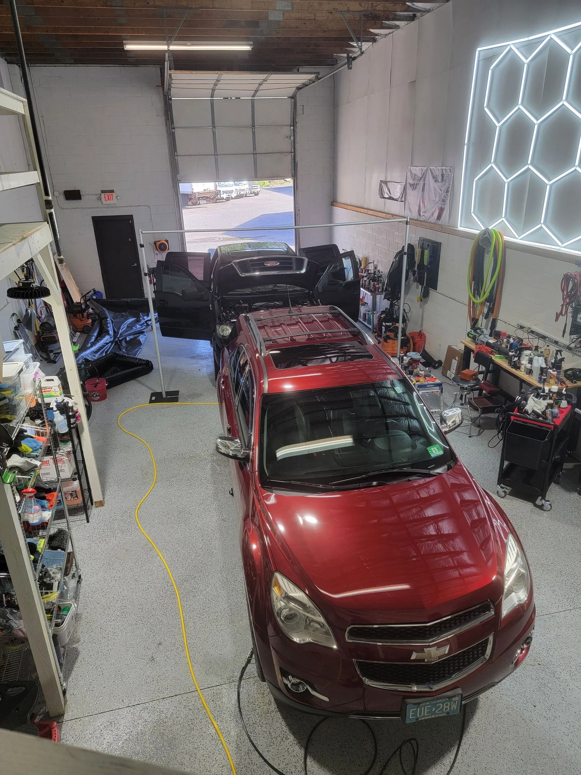A red SUV parked in a garage with a hexagonal LED light wall, specialized equipment, and open doors for detailing.