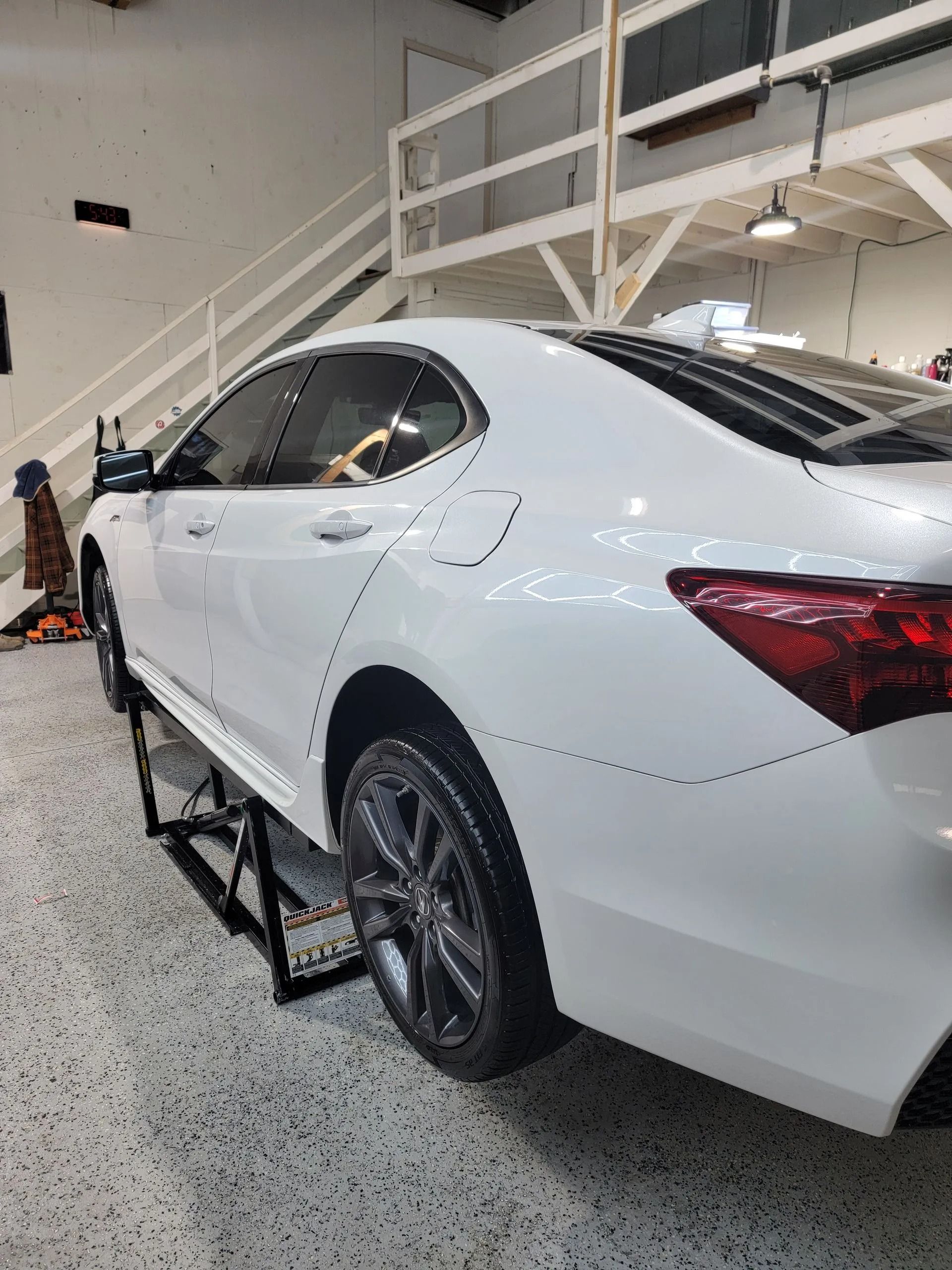 A white sedan elevated on a mechanic's lift inside a brightly lit garage with a white staircase in the background.