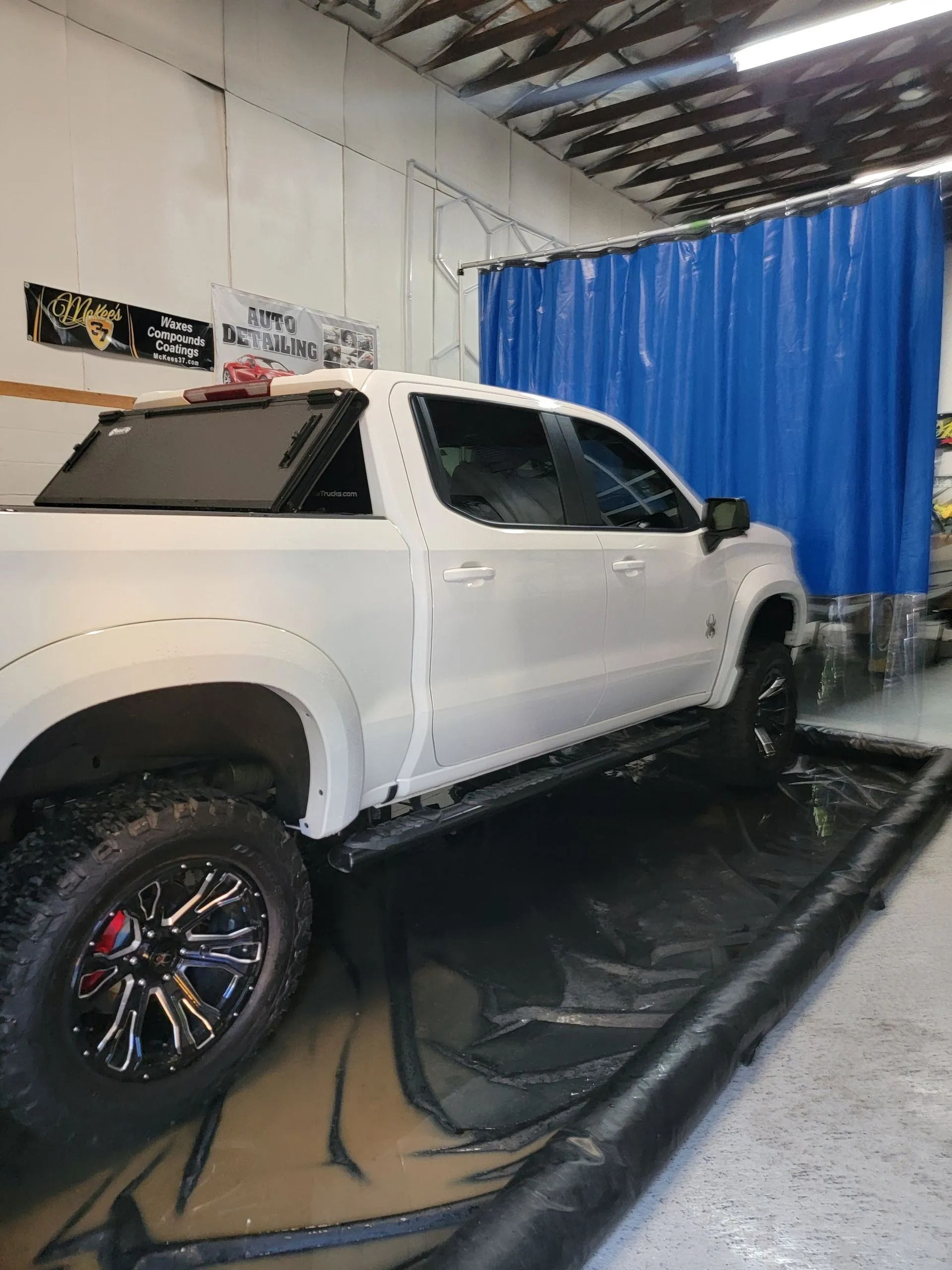 A white pickup truck sits in a containment mat inside a garage, with a blue curtain visible in the background.