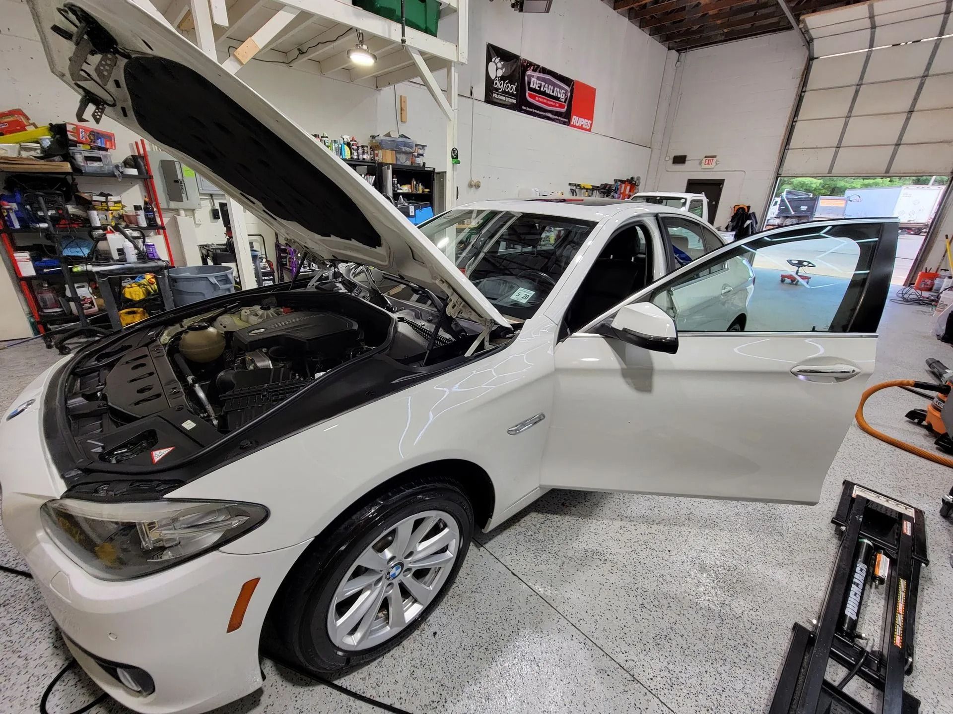 A white car with its hood and driver-side door open, parked inside a brightly lit automotive workshop.