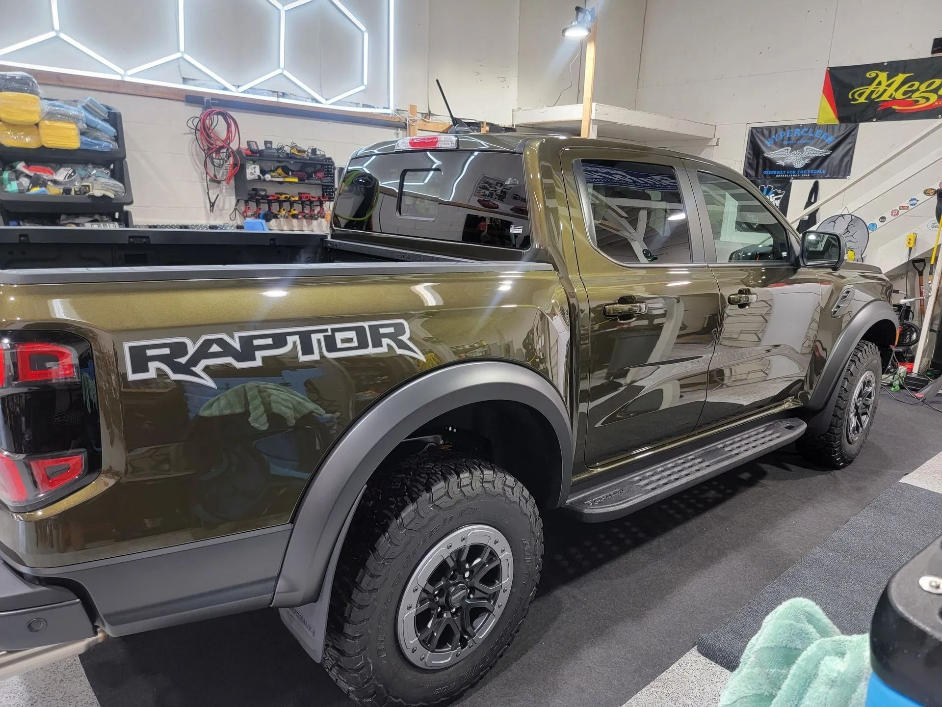 An olive green Ford Ranger Raptor pickup truck parked in a well-lit garage with automotive detailing supplies.