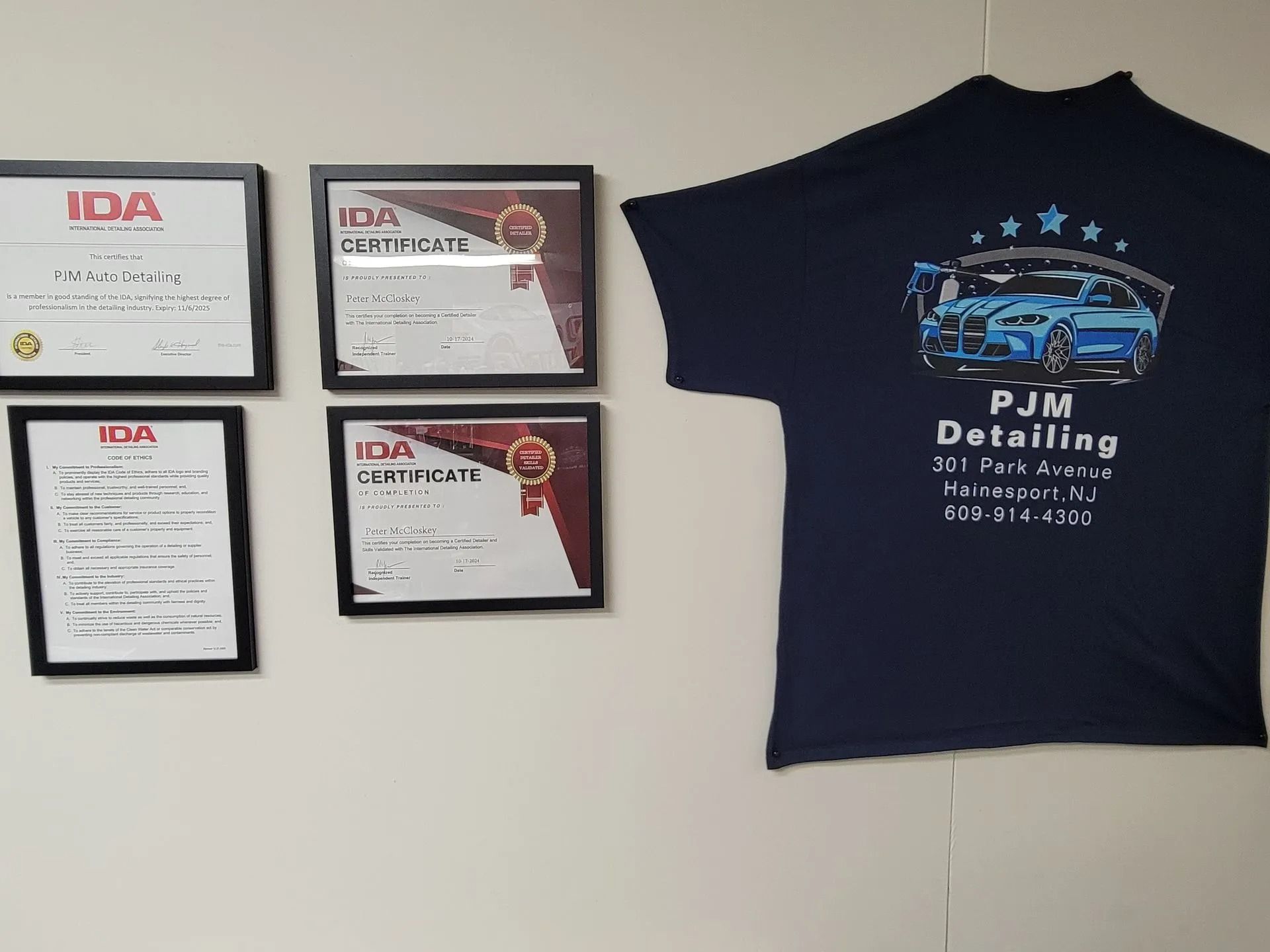 Four IDA certificates framed on a light wall next to a navy blue PJM Detailing t-shirt featuring a blue car graphic.