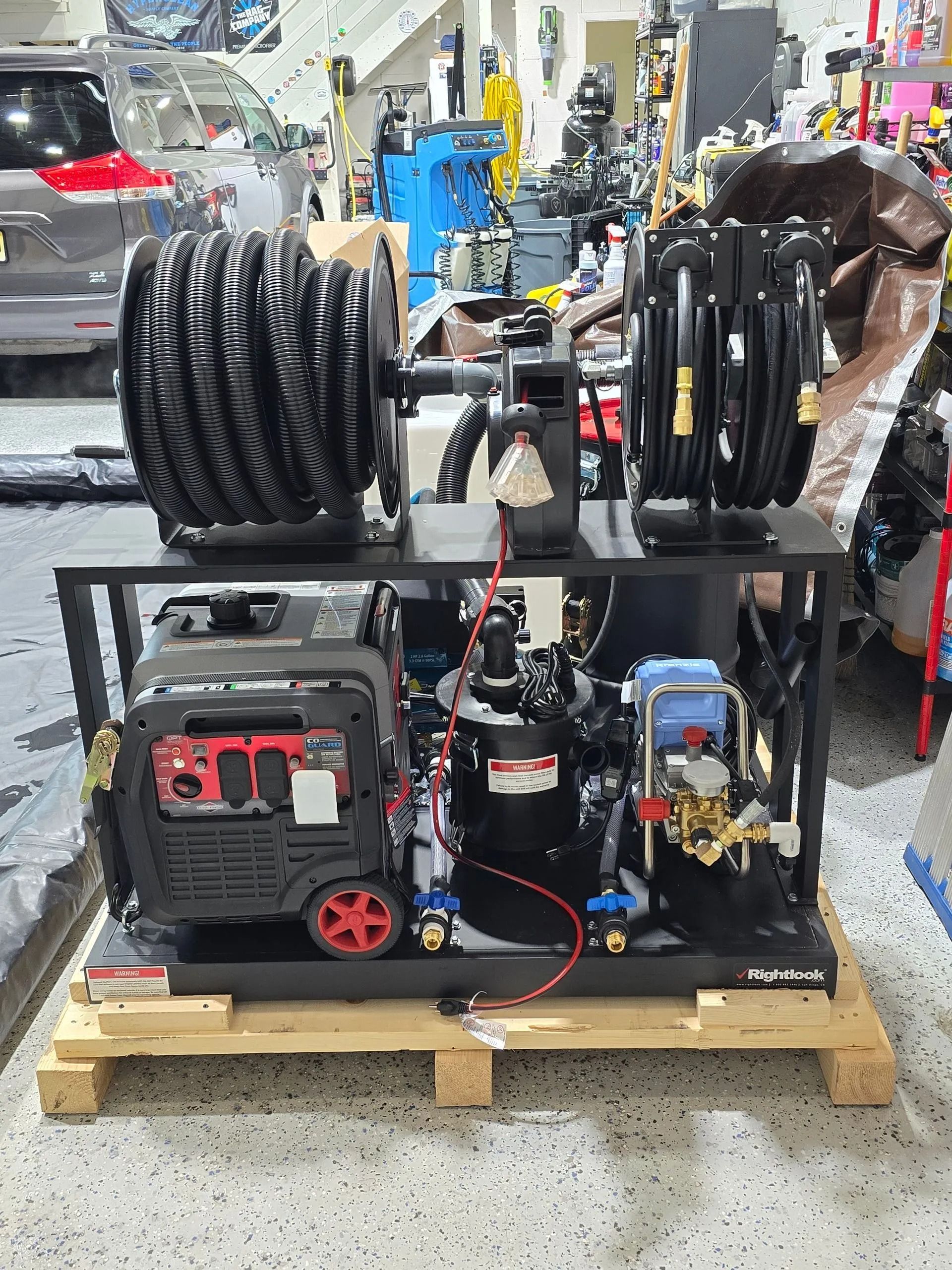 A black industrial pressure washing system on a wooden pallet in a garage, featuring two hose reels and a red generator.