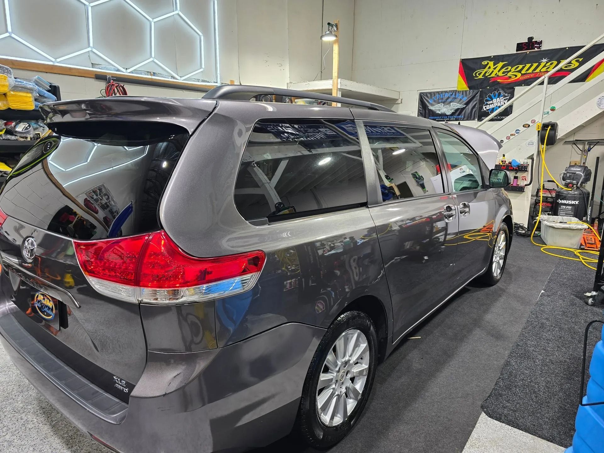 A gray minivan parked inside an auto detailing shop with bright lighting and workshop equipment in the background.
