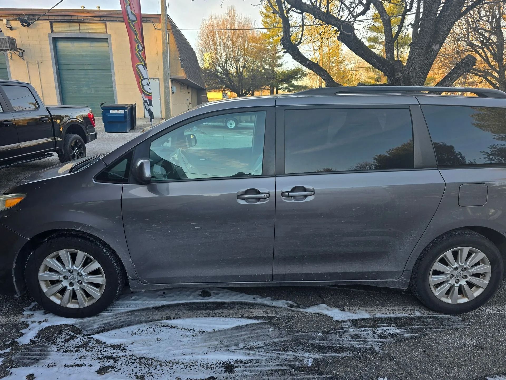 A dark gray minivan parked outdoors on a pavement with some light snow, with a truck visible in the background.