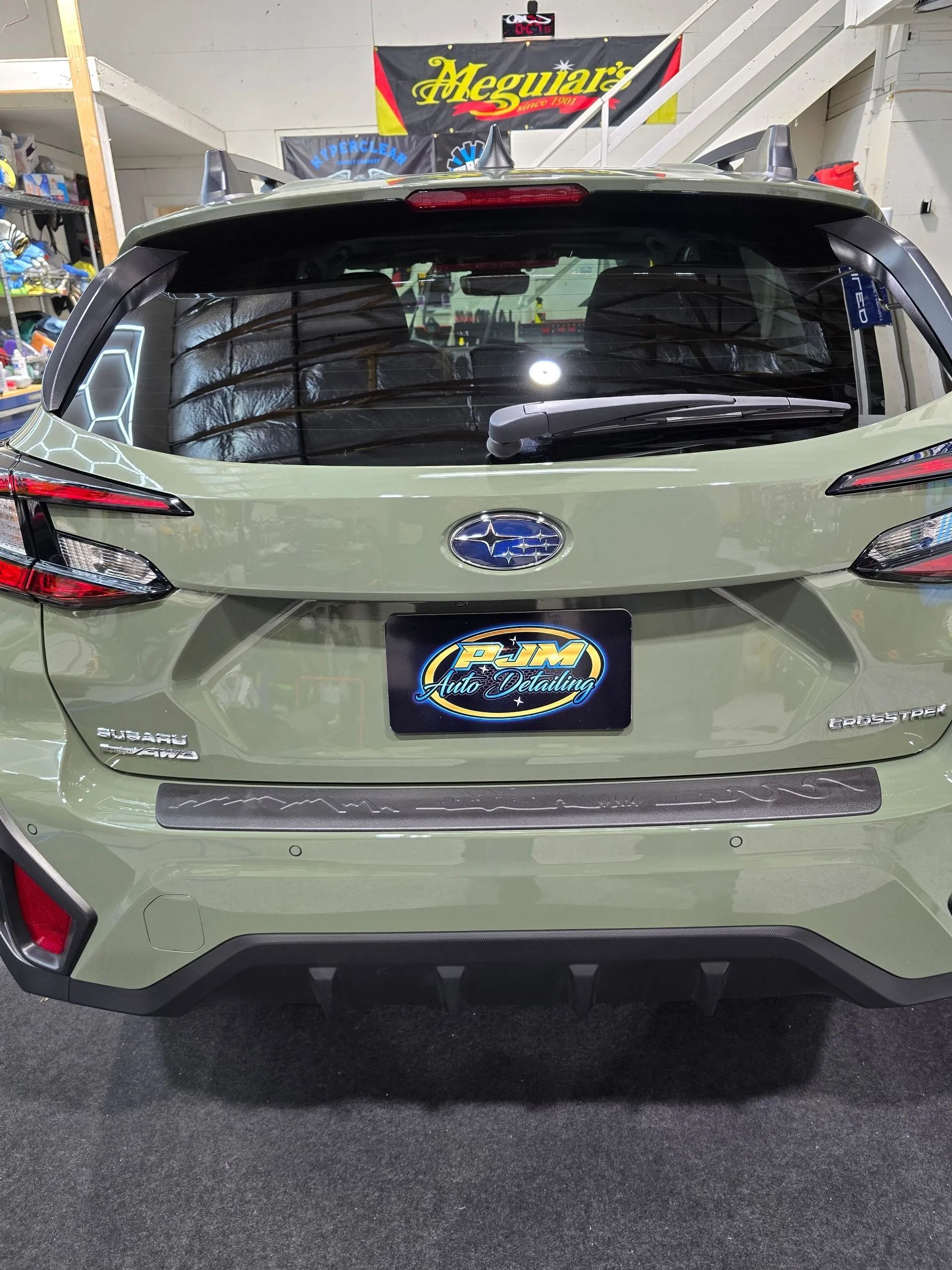 Back view of a sage green Subaru Crosstrek parked inside an auto shop with a Meguiar’s banner in the background.