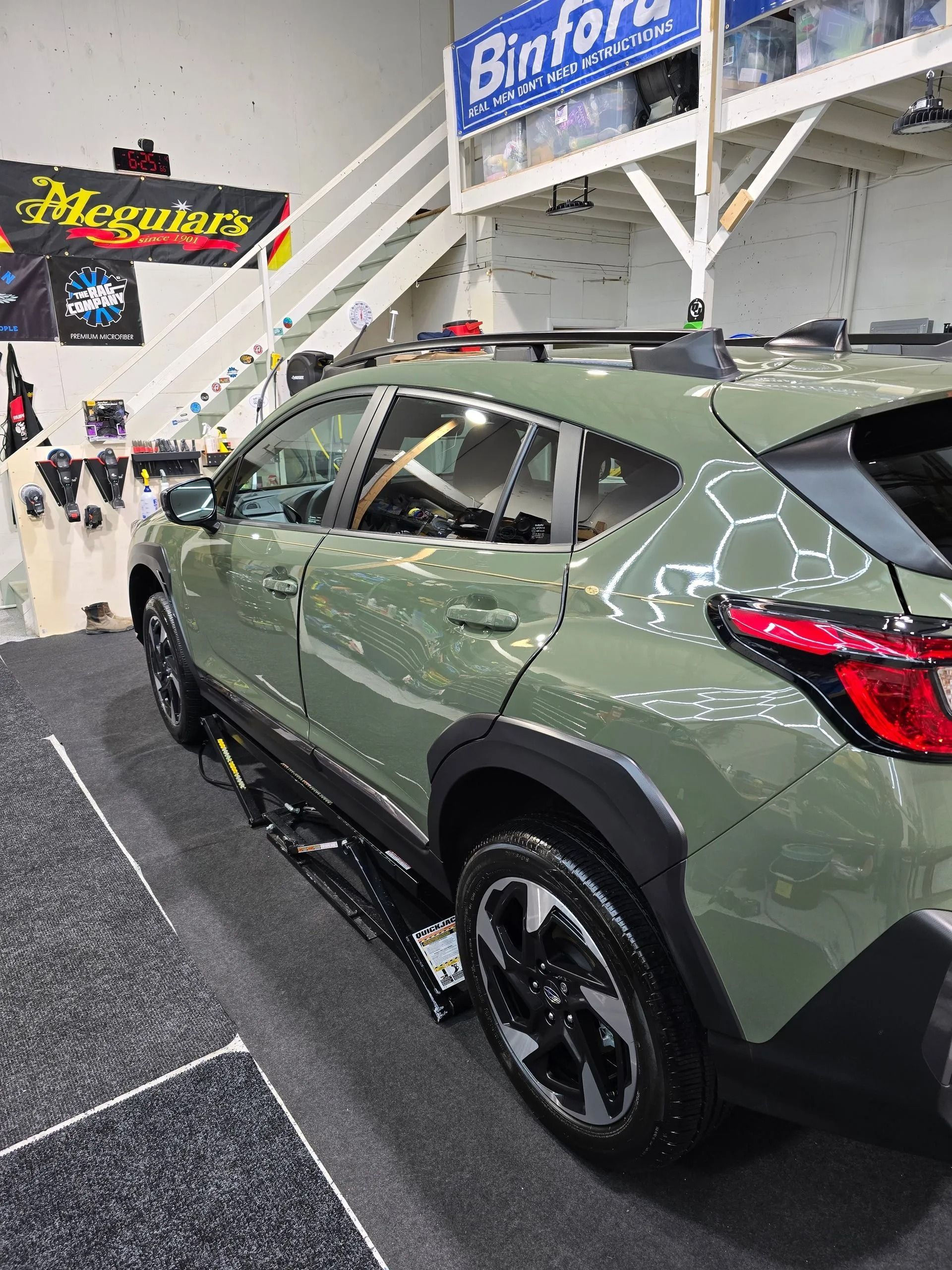 A light green Subaru Crosstrek parked on a lift inside a garage with white stairs and branded wall signs.