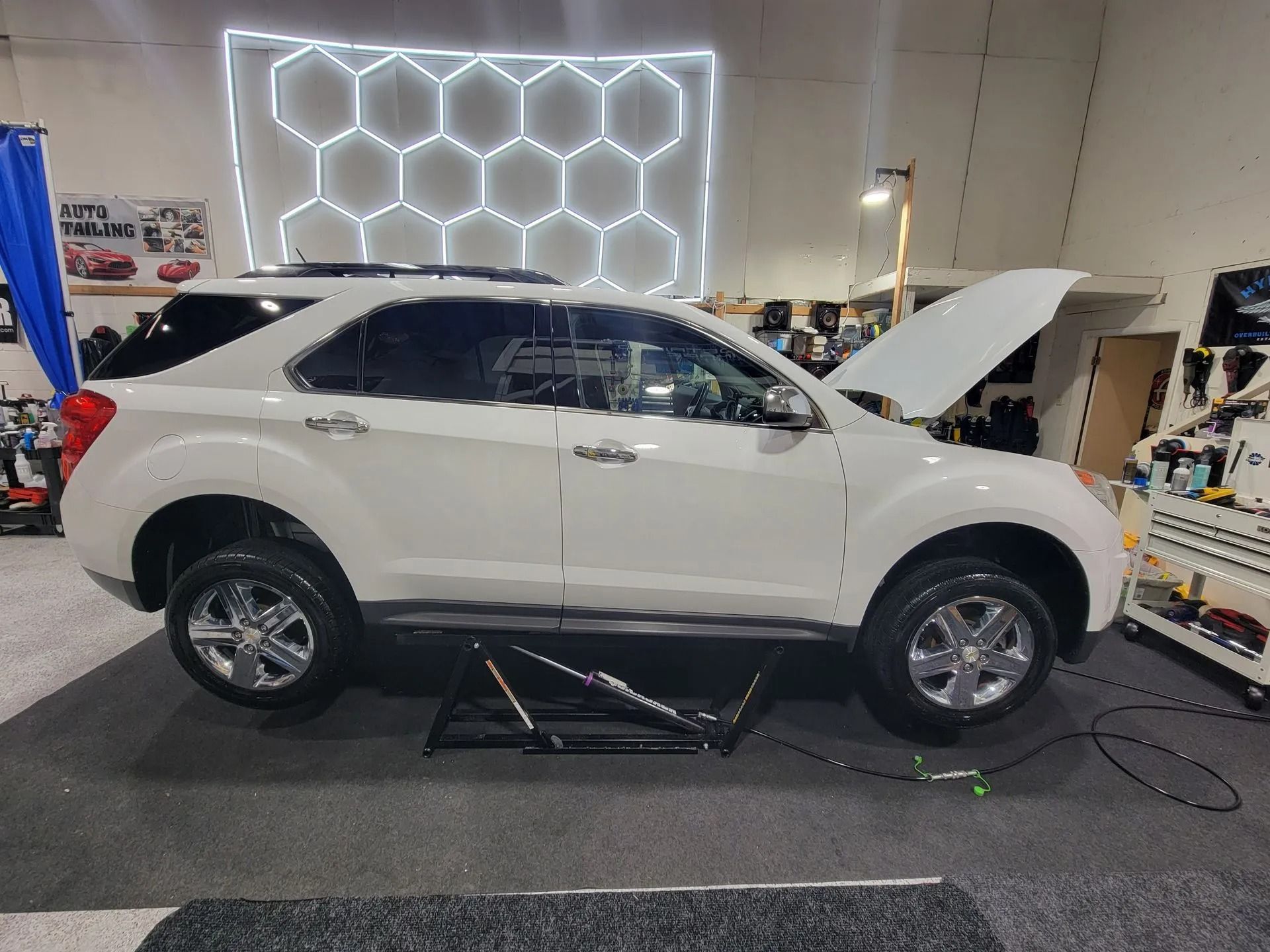 A white SUV with its hood raised is being lifted by a low-profile car jack inside an automotive repair shop.