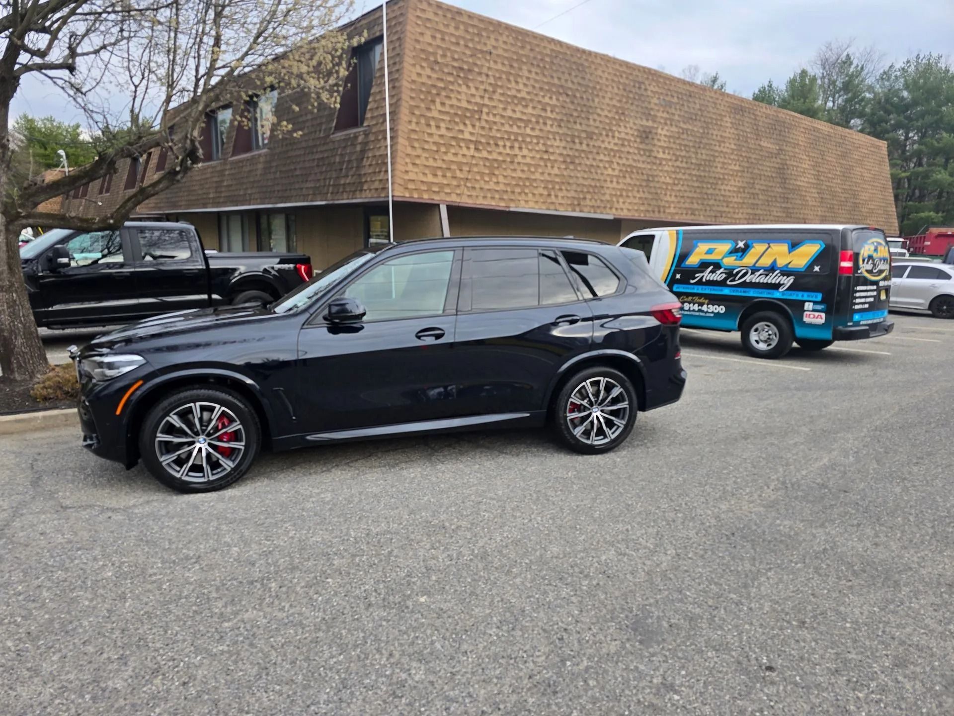 A black SUV parked in an asphalt lot next to a PJM Auto Detailing van and a black truck, with a brown building in back.