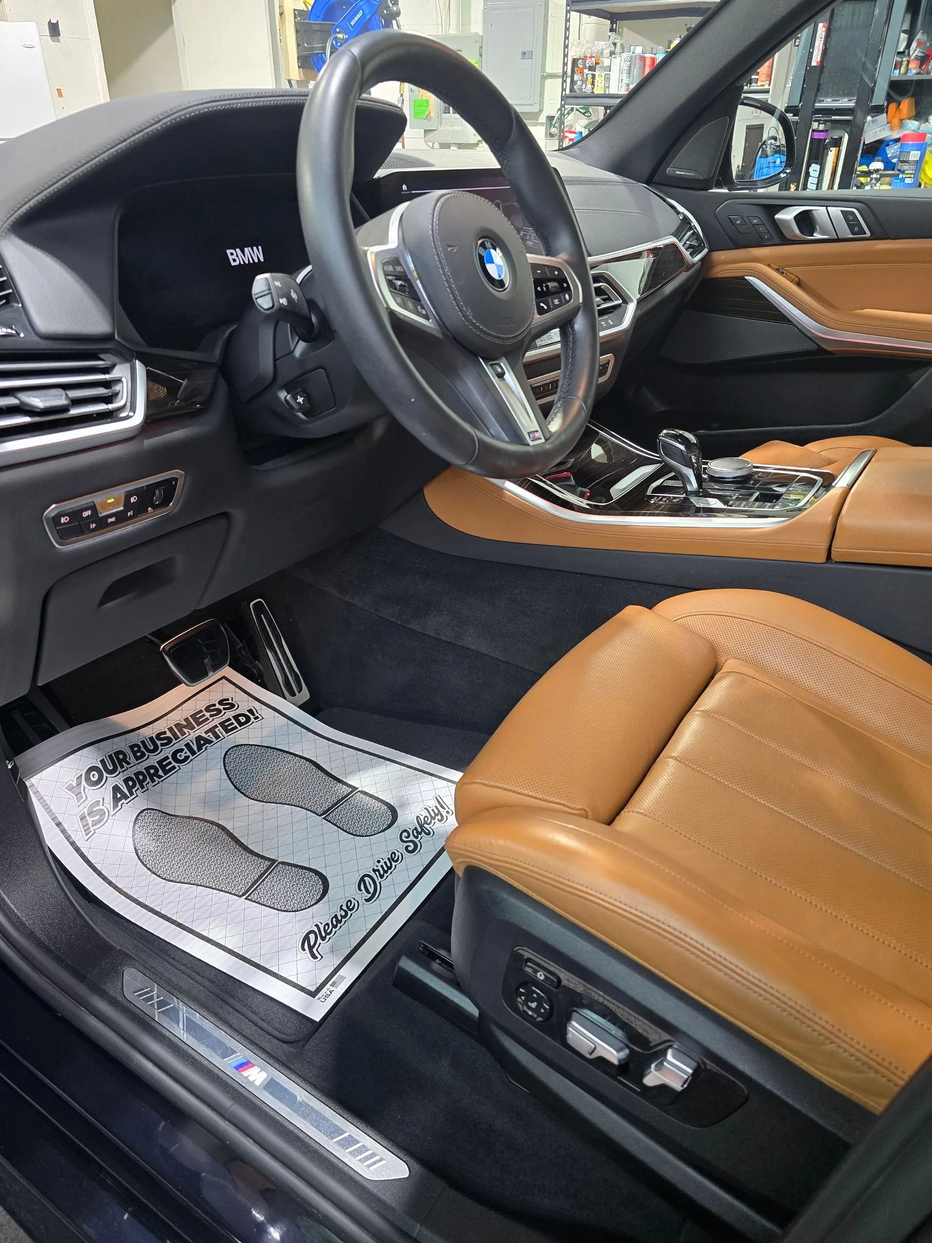 A luxury car interior features tan leather seats, a black dashboard, a leather-wrapped steering wheel, and floor liners.