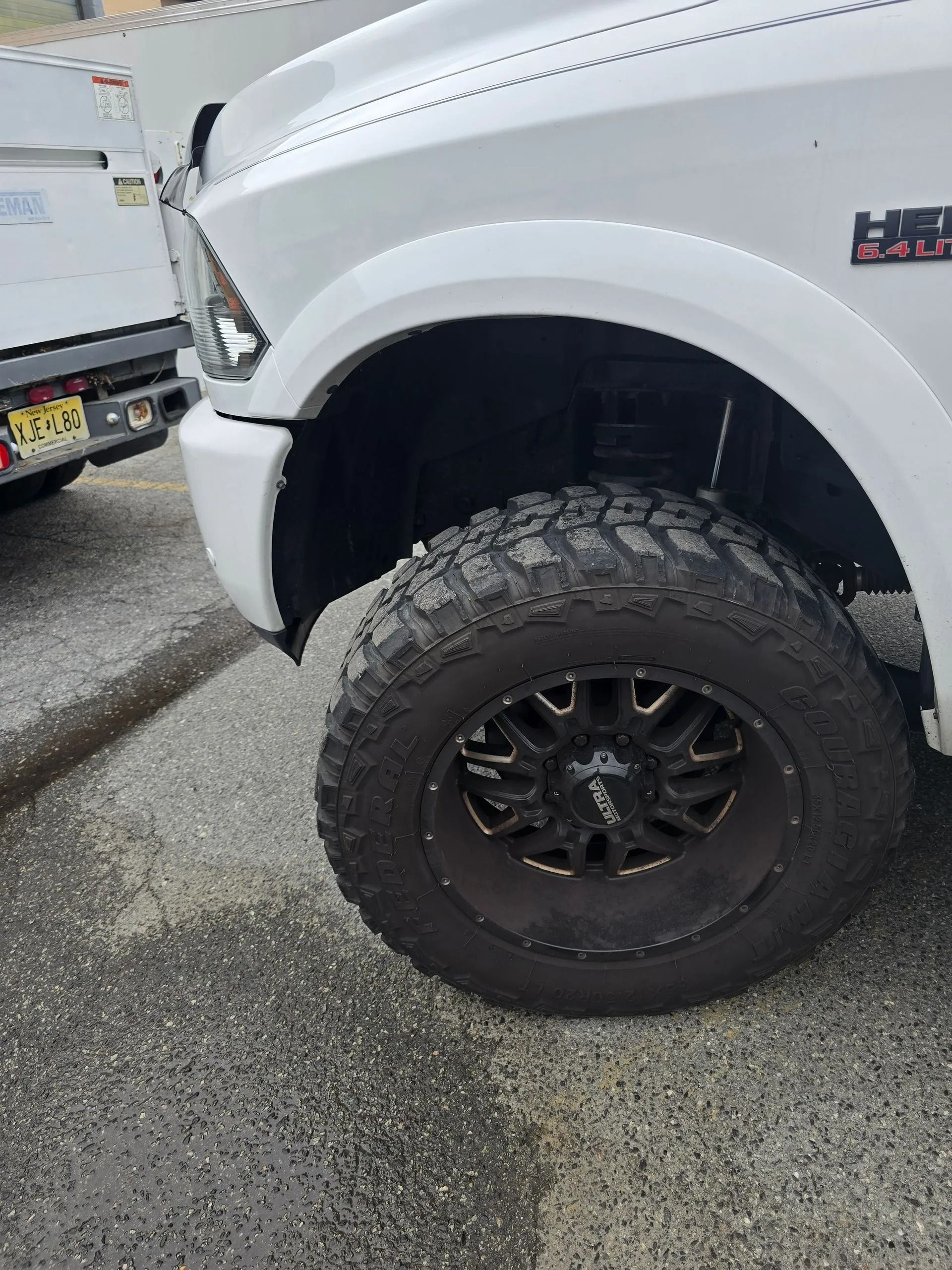 Close-up of a white Ram truck's front wheel, showing a large off-road tire on a dark custom rim, parked on pavement.