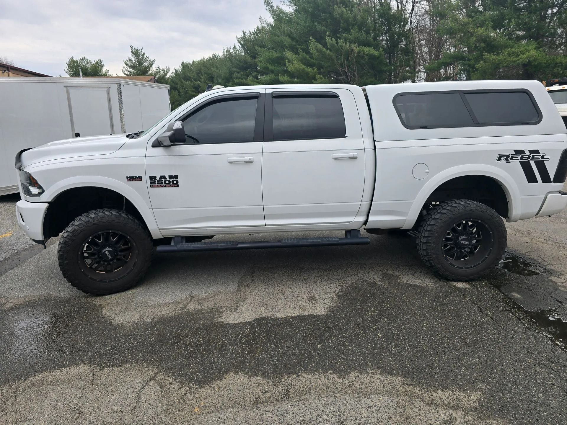 A white Ram pickup truck with a camper shell and aftermarket black wheels parked on an asphalt surface.