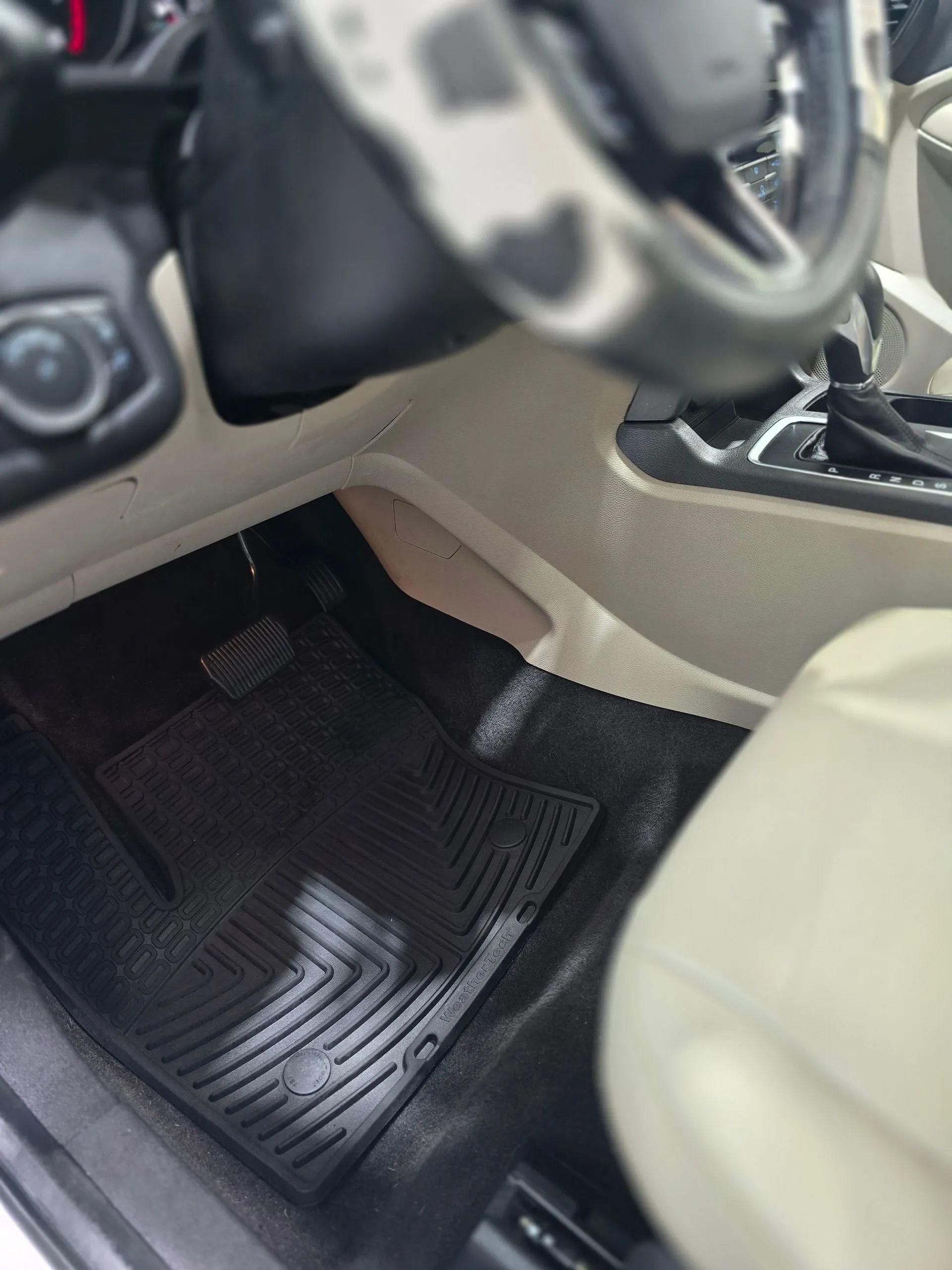 An interior view of a car’s front footwell, showing black rubber floor mats, a beige interior trim, and the steering wheel.