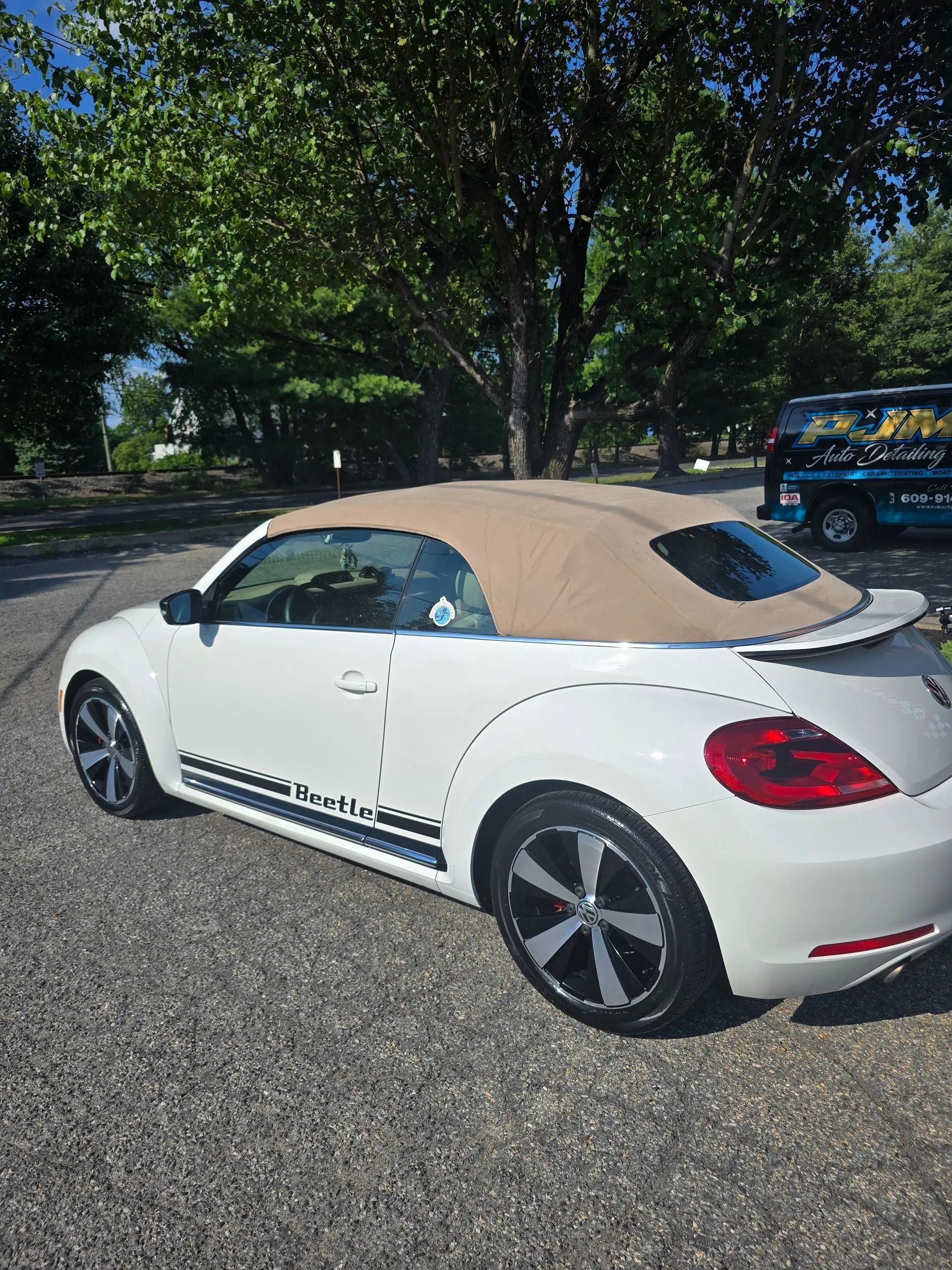 A white Volkswagen Beetle convertible with a tan soft top and custom side decals parked on a gravel lot.