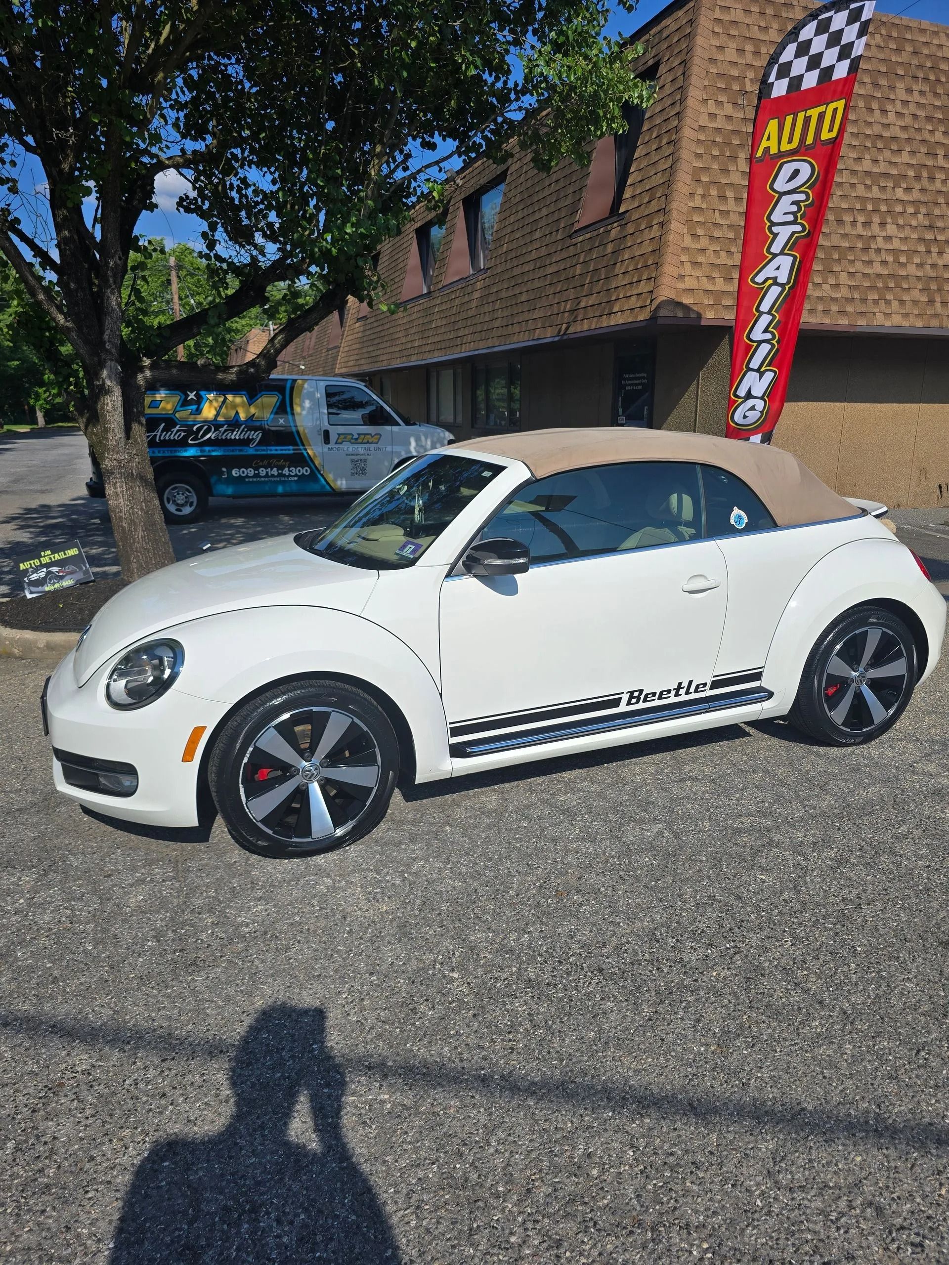 A white Volkswagen Beetle convertible parked in a lot in front of a brick building with an auto detailing banner.