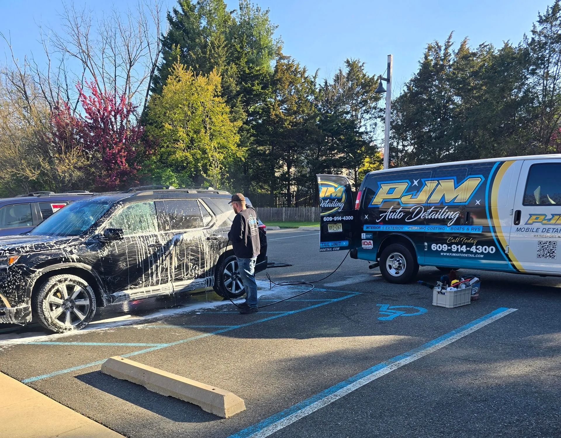 A person soaps a black SUV in a parking lot next to a PJM Auto Detailing van.