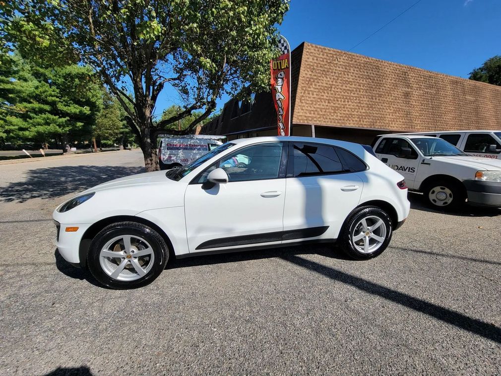 A white Porsche SUV parked on a gravel lot in front of a building with a textured brown facade under a blue sky.