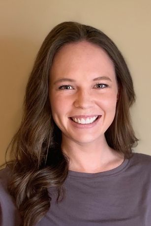 Smiling woman with long brown hair against a beige background, wearing a gray shirt.