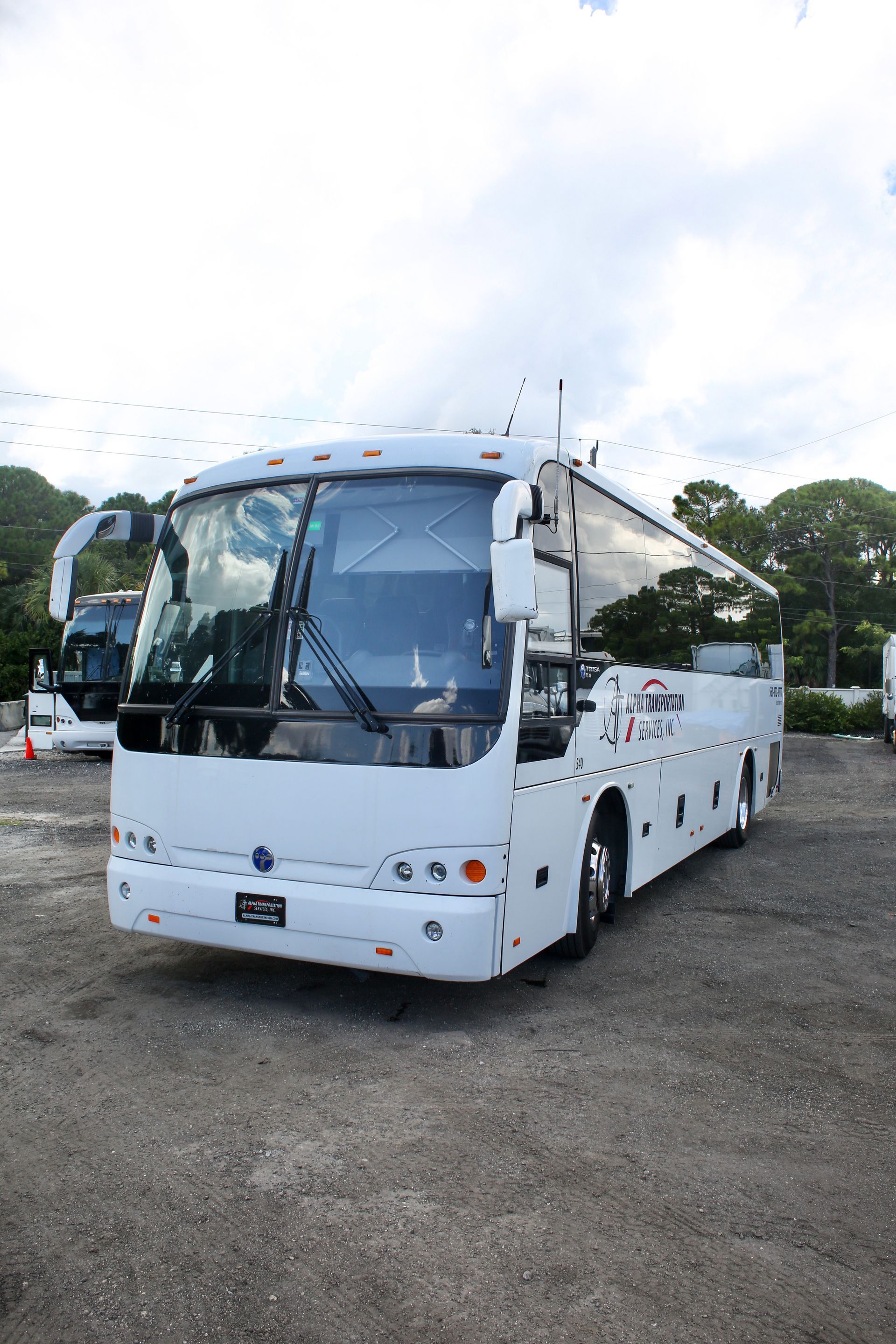 White coach bus parked on gravel under a cloudy sky, front view with large tinted windshield.