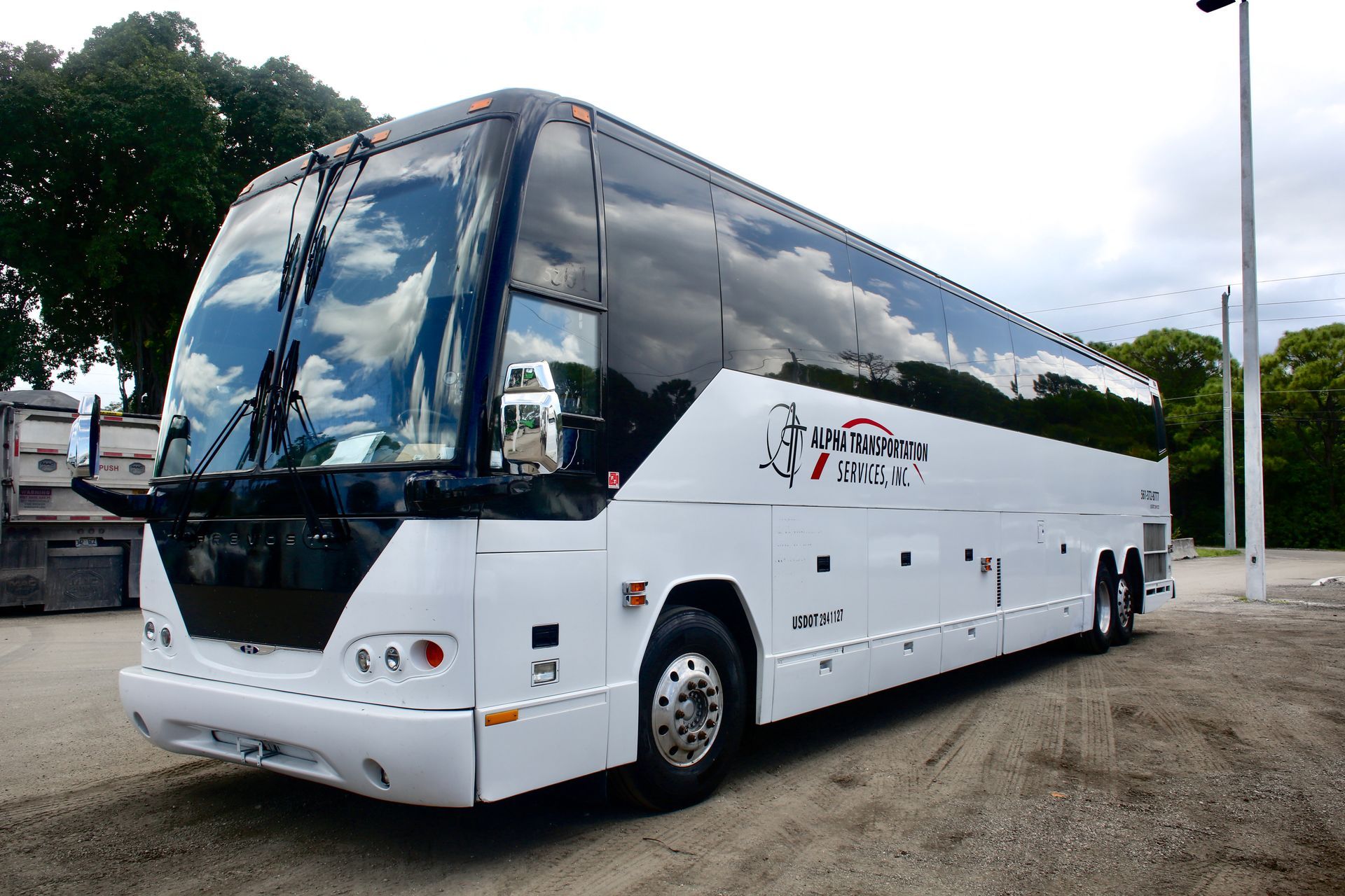 White coach bus parked outdoors on gravel, with dark tinted windows and trees in the background