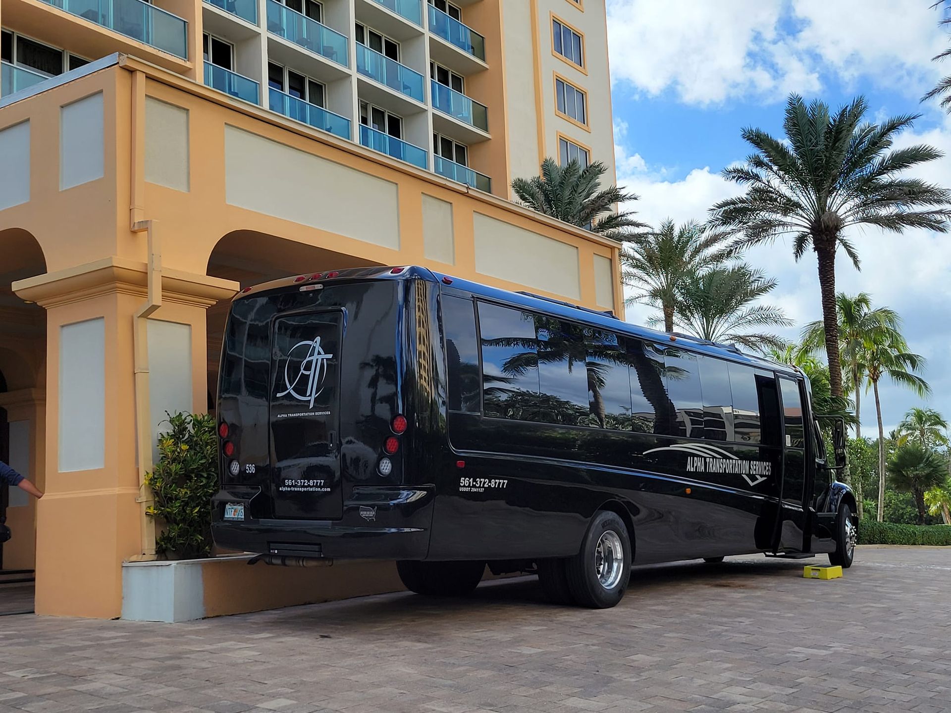 Black shuttle bus parked outside a beige hotel with palm trees on a sunny day