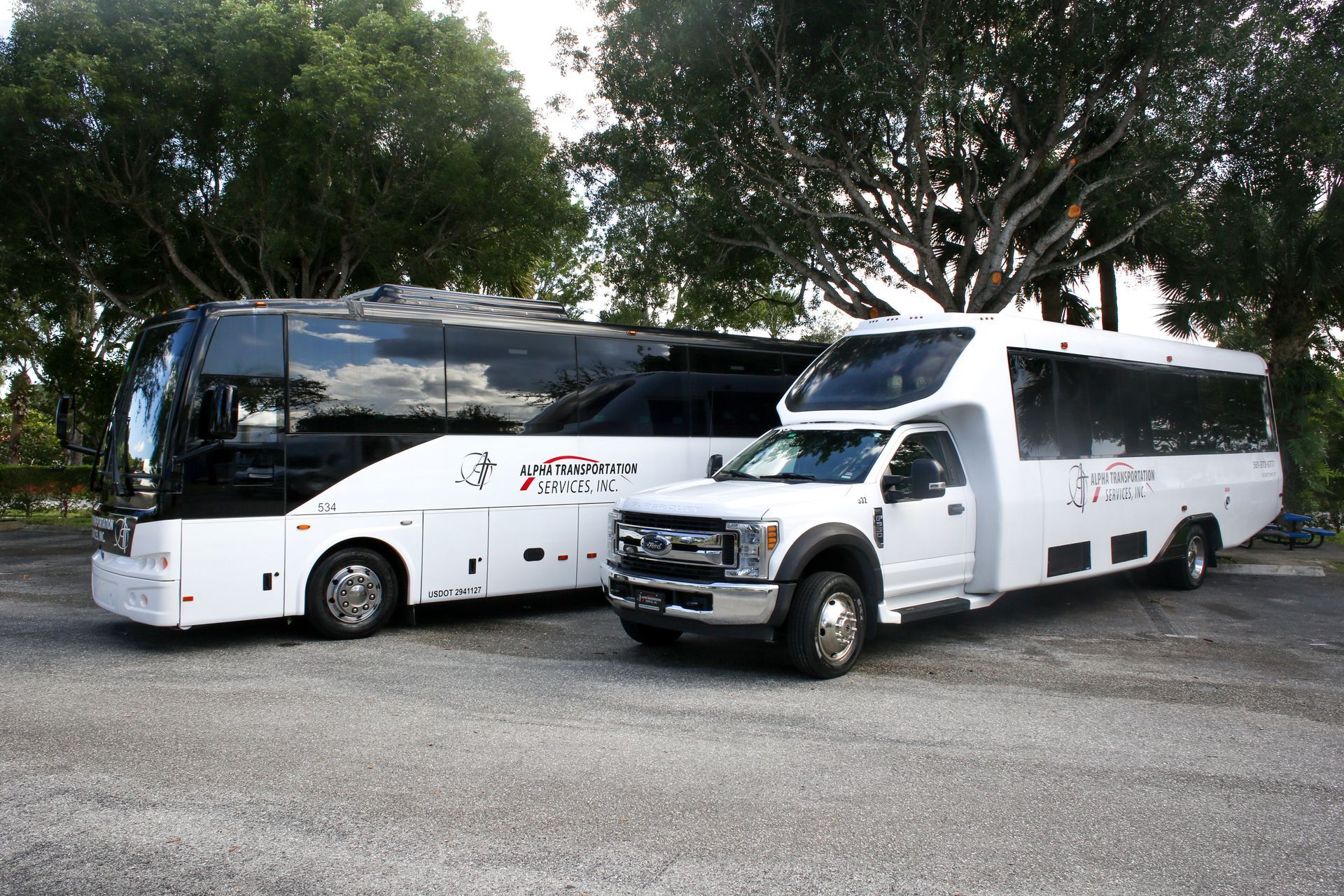 White tour bus and white van parked side by side on gravel under trees