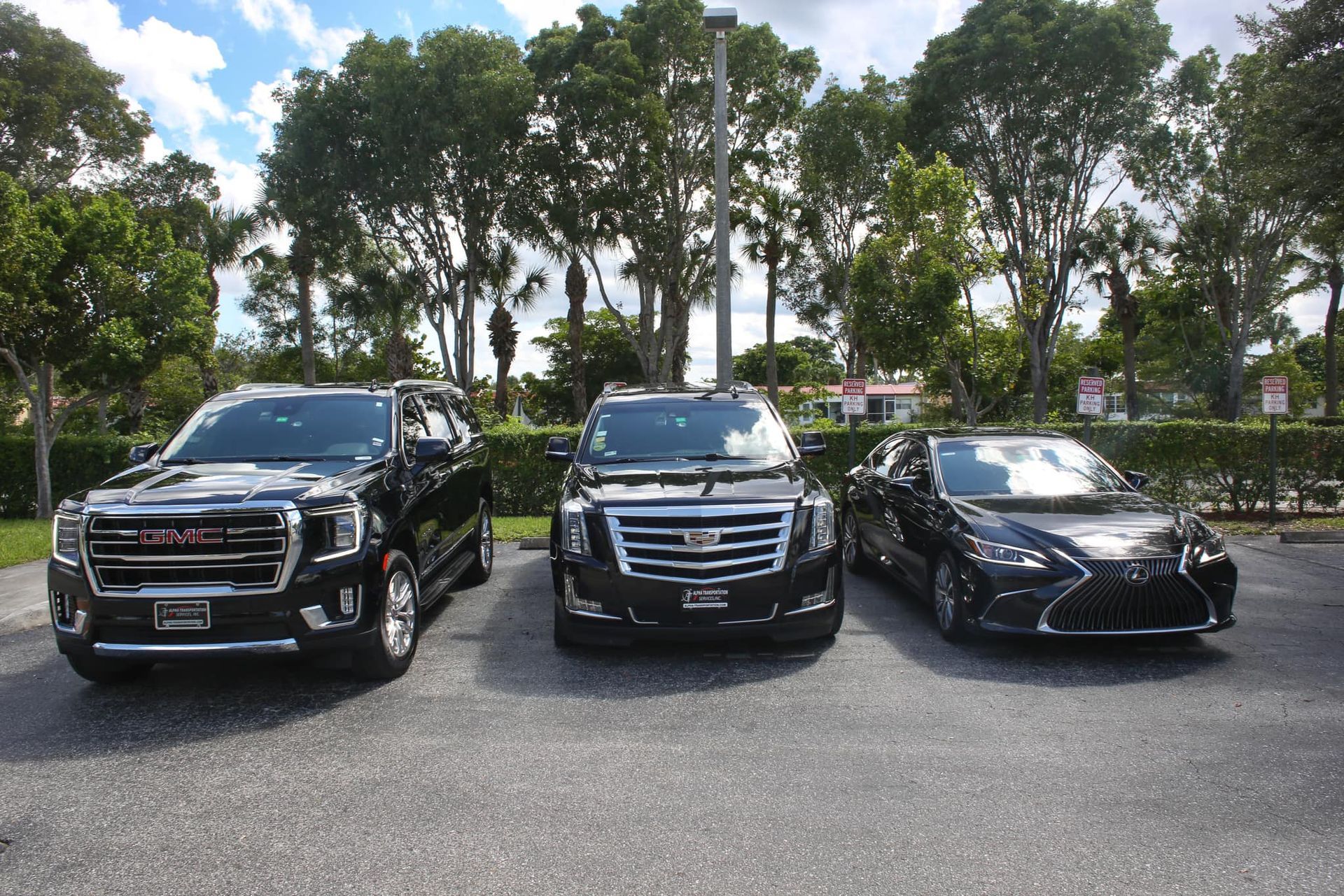 Three parked cars on a lot, including two black SUVs and a black sedan, with trees in the background.