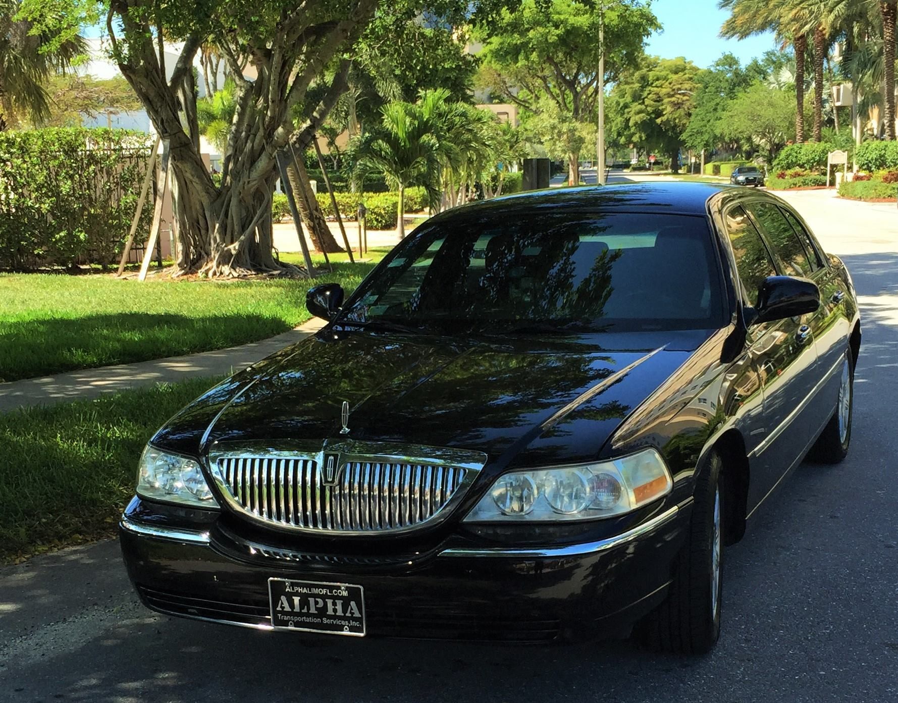 Black sedan parked on a tree-lined residential street in bright sunlight