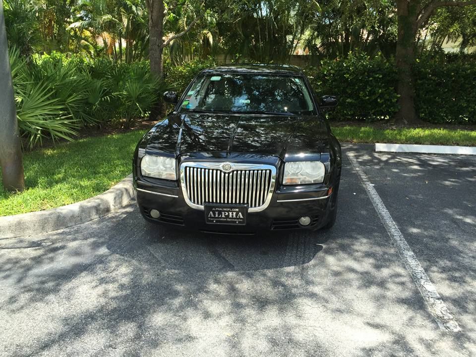 Black sedan parked on a sunlit driveway beside lush green trees and shrubs.