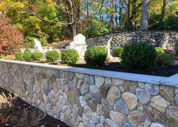 A stone wall surrounds a garden with chairs and bushes.