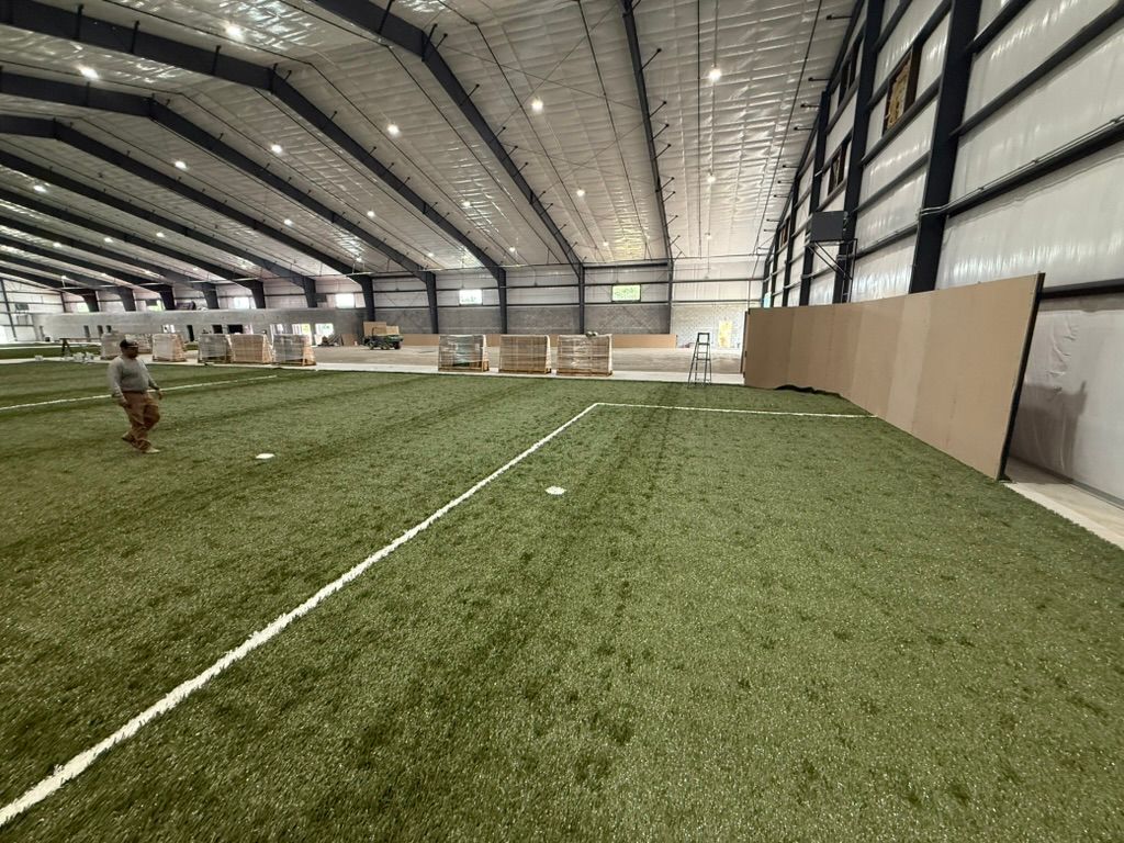 An indoor artificial turf field inside a large metal building. A person walks on the field near a white line.
