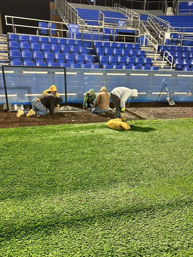 People working on a baseball field. They are bent over, possibly planting or tending to flowers along the infield. Blue stadium seating in the background.