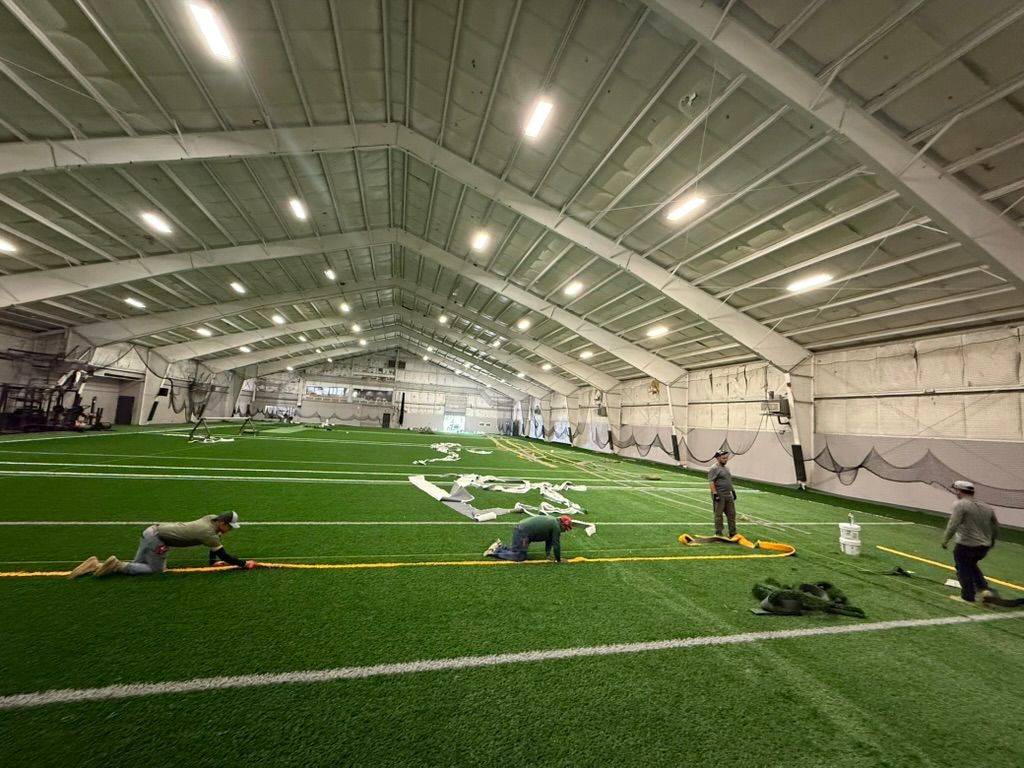 Indoor athletic field with artificial turf, people working, and bright overhead lights.