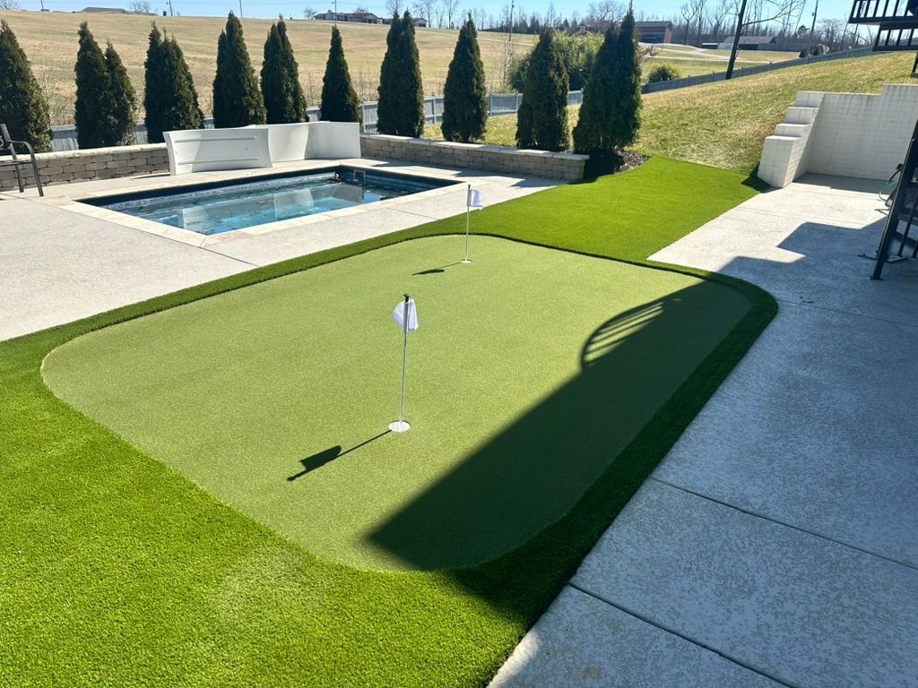 Backyard with putting green, pool, and small trees. Green grass and artificial turf. Sunny day.