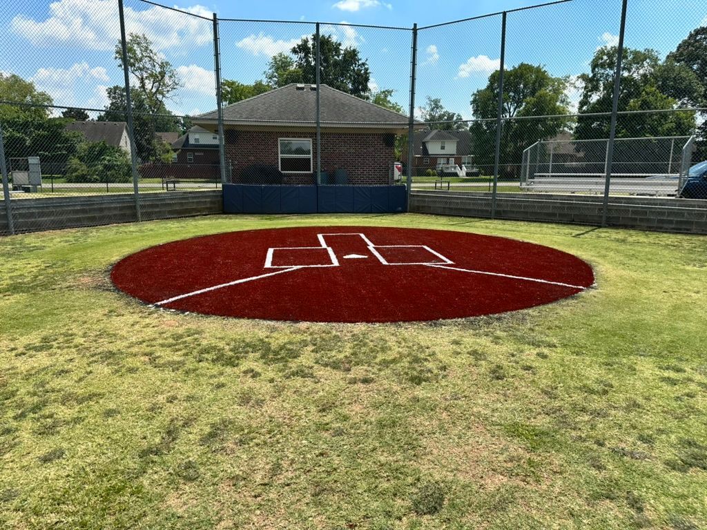 Baseball practice area with red infield, white lines, protective net, and a small building in the background.