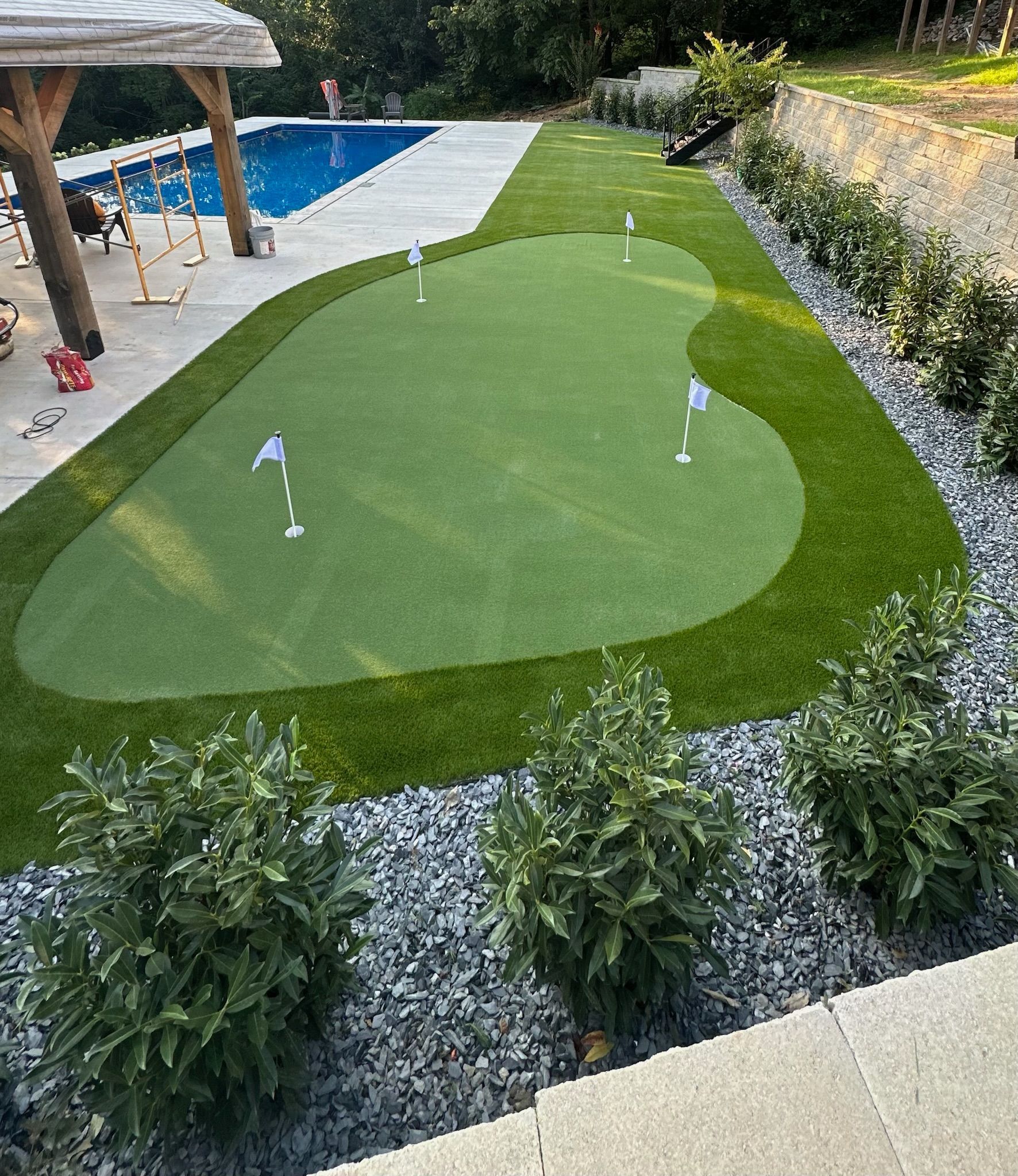 Backyard putting green next to a pool. Lush green turf with flags, surrounded by landscaping, and a patio.
