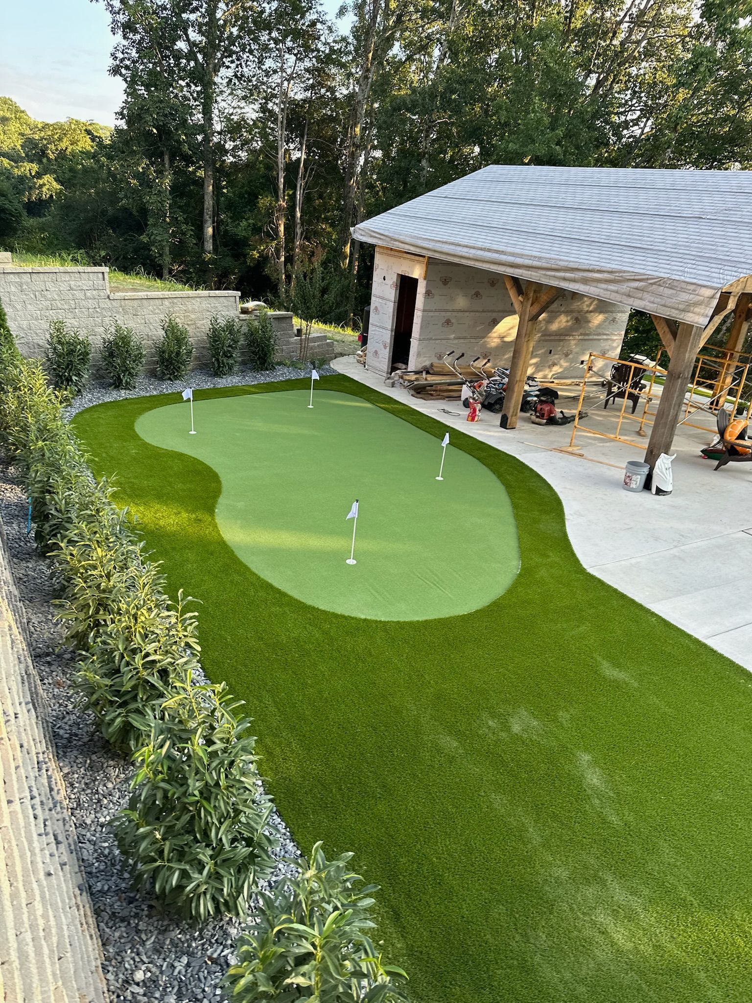 A putting green with multiple holes, surrounded by a lush lawn and trimmed shrubs, beside a stone structure with an open-air roof.