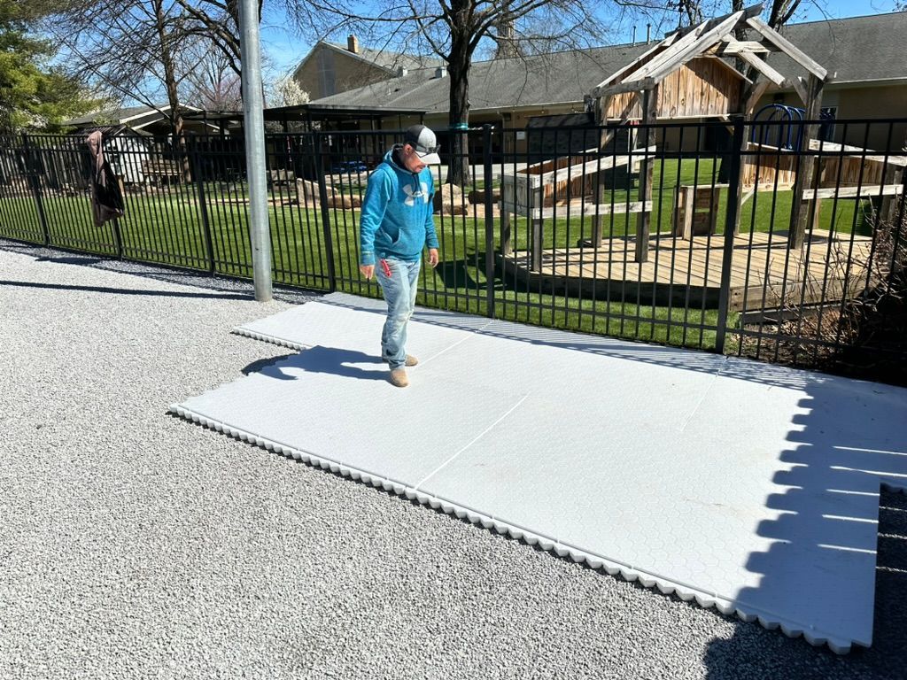 Person standing on white mats, gravel ground, black fence, and building in the background.