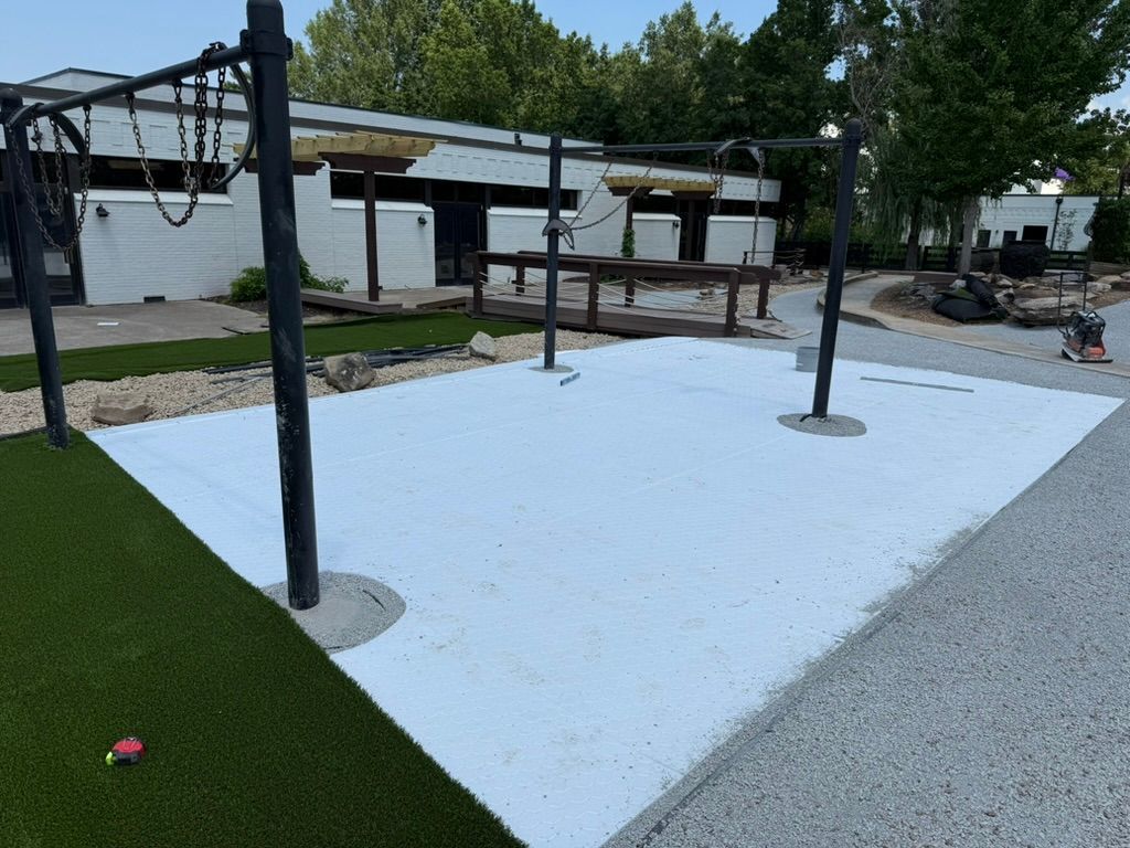 A light-colored concrete patio with black metal structures, artificial turf, and gravel, outside a building.