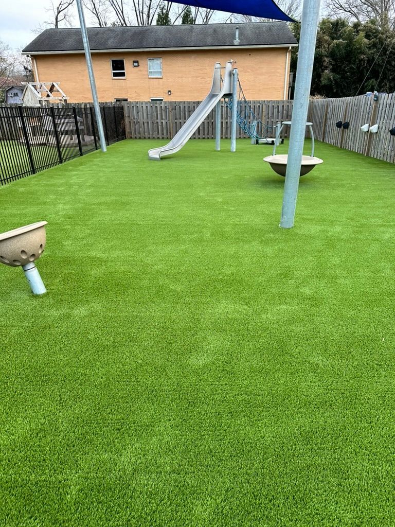 Playground with green turf, a slide, and play equipment, enclosed by a fence.