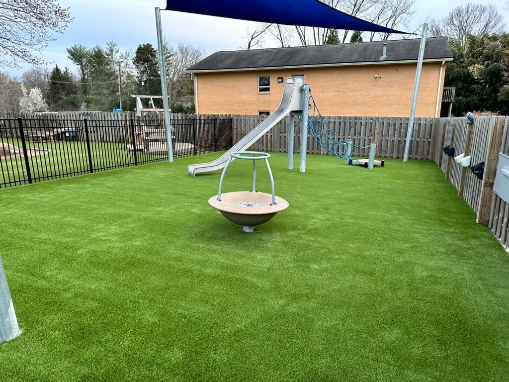 Playground with artificial turf, slide, and spinner, enclosed by black and wooden fences, under a shade sail.