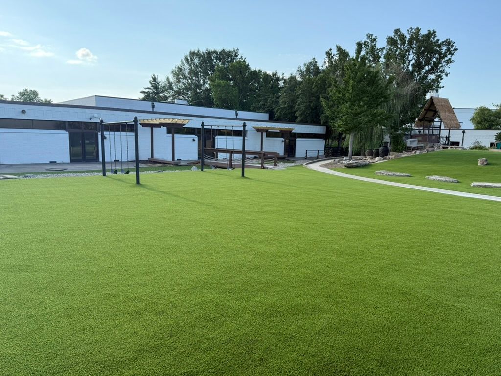Exterior of a building with artificial turf. Pergola and play structure in the foreground. Trees and blue sky.