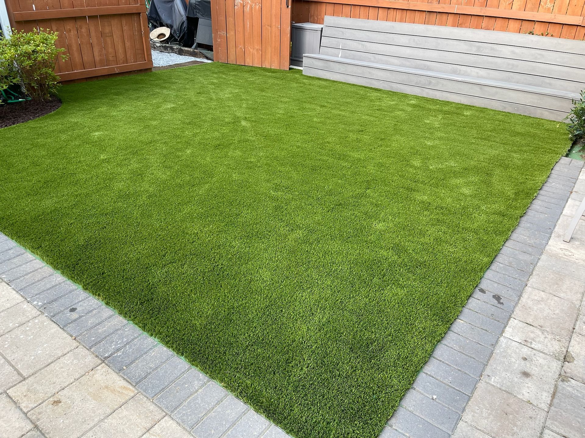 Green artificial turf in a small fenced yard with brick border and wooden fence.