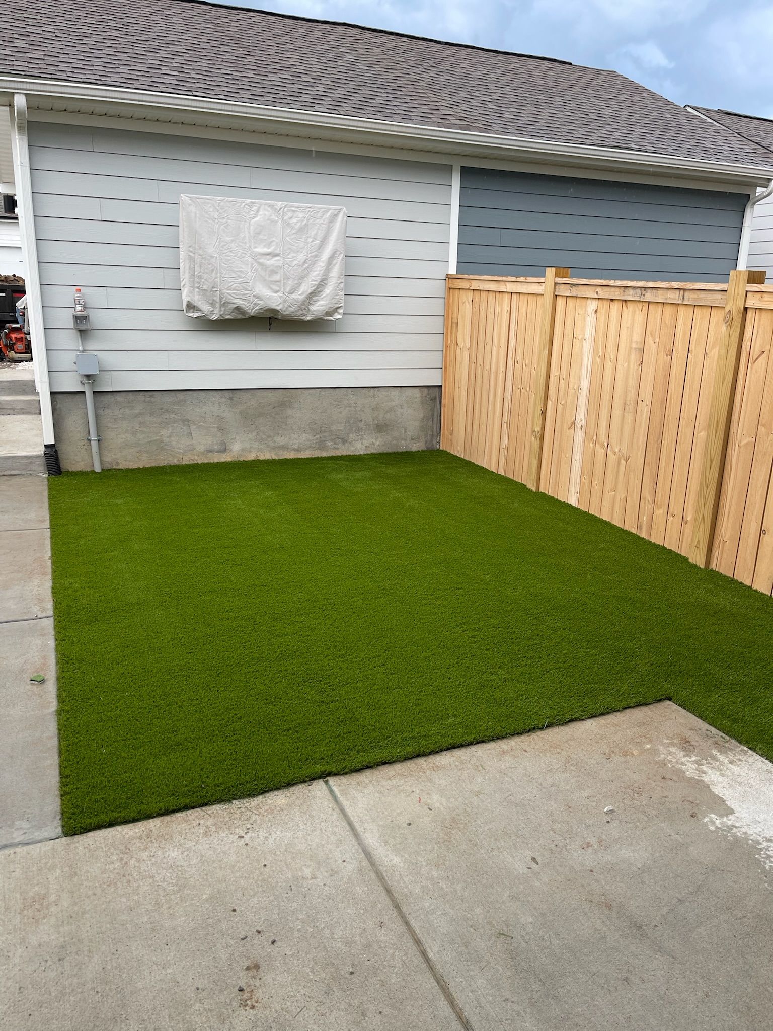 Small backyard with green artificial turf, light gray house, wooden fence, and concrete.