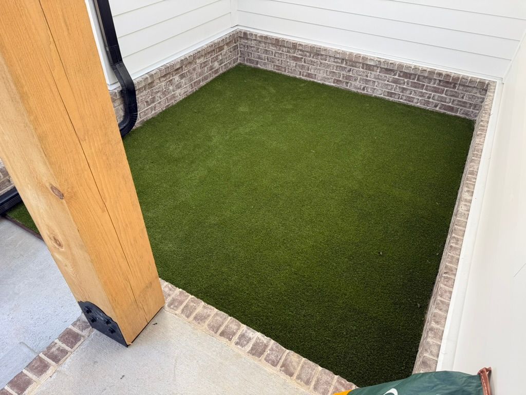 Small, square area with artificial turf, brick edging, and white walls. A wooden beam is on the left.