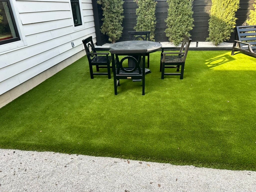 A small backyard with artificial green grass, black table, and two chairs. Gravel border next to white house.