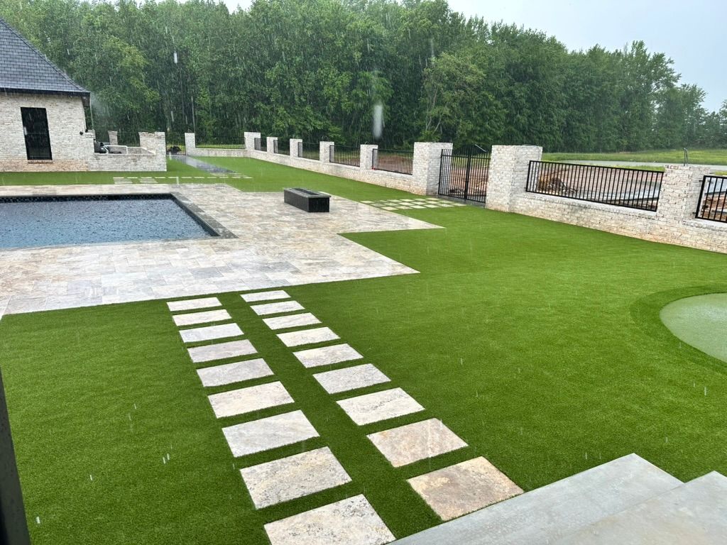 Stone patio with pool, stepping stones through green turf, stone walls, and trees. Overcast weather.
