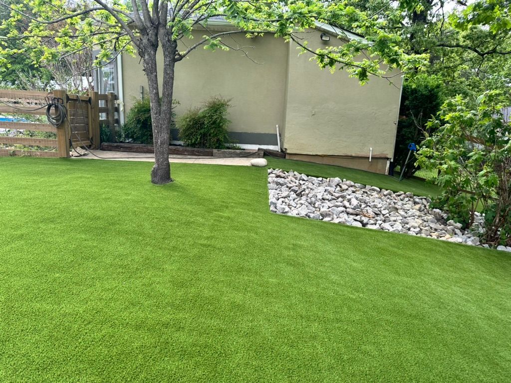 Green artificial turf in a backyard, with a tree, stone border, and a building.