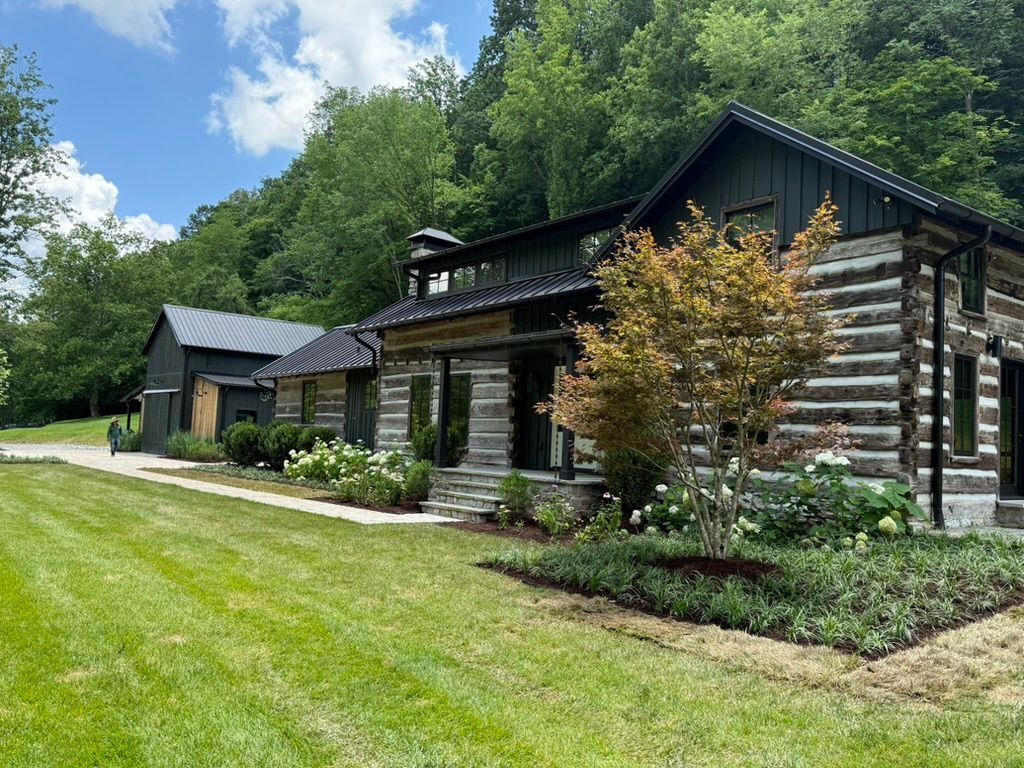 Log cabin with a dark roof and siding, surrounded by a green lawn and trees.