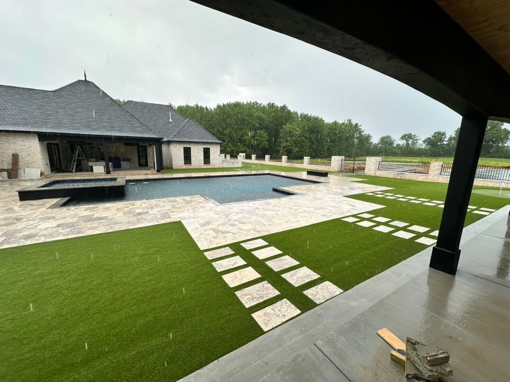 Backyard with pool, stone patio, green turf, and a house under a cloudy sky.